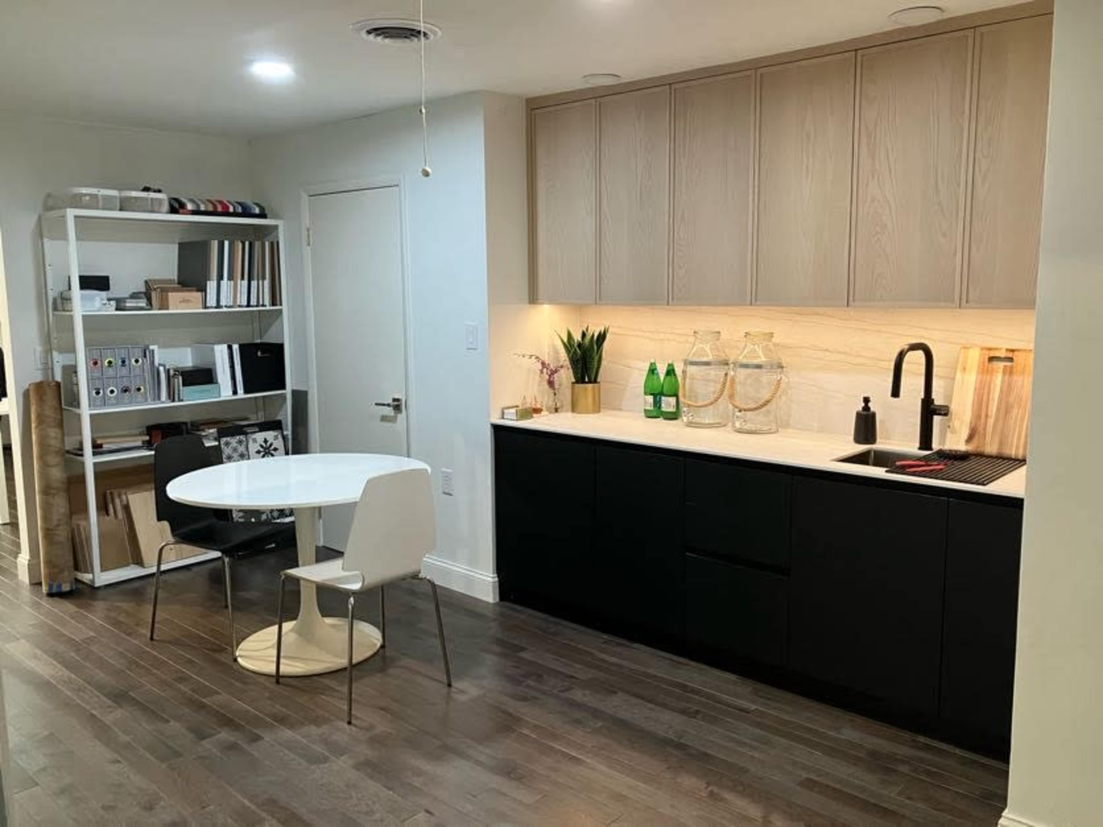 A modern kitchen featuring a black and white color scheme, with a round table and chairs, a shelf of organized items, and a countertop with bottles and decorative vases.