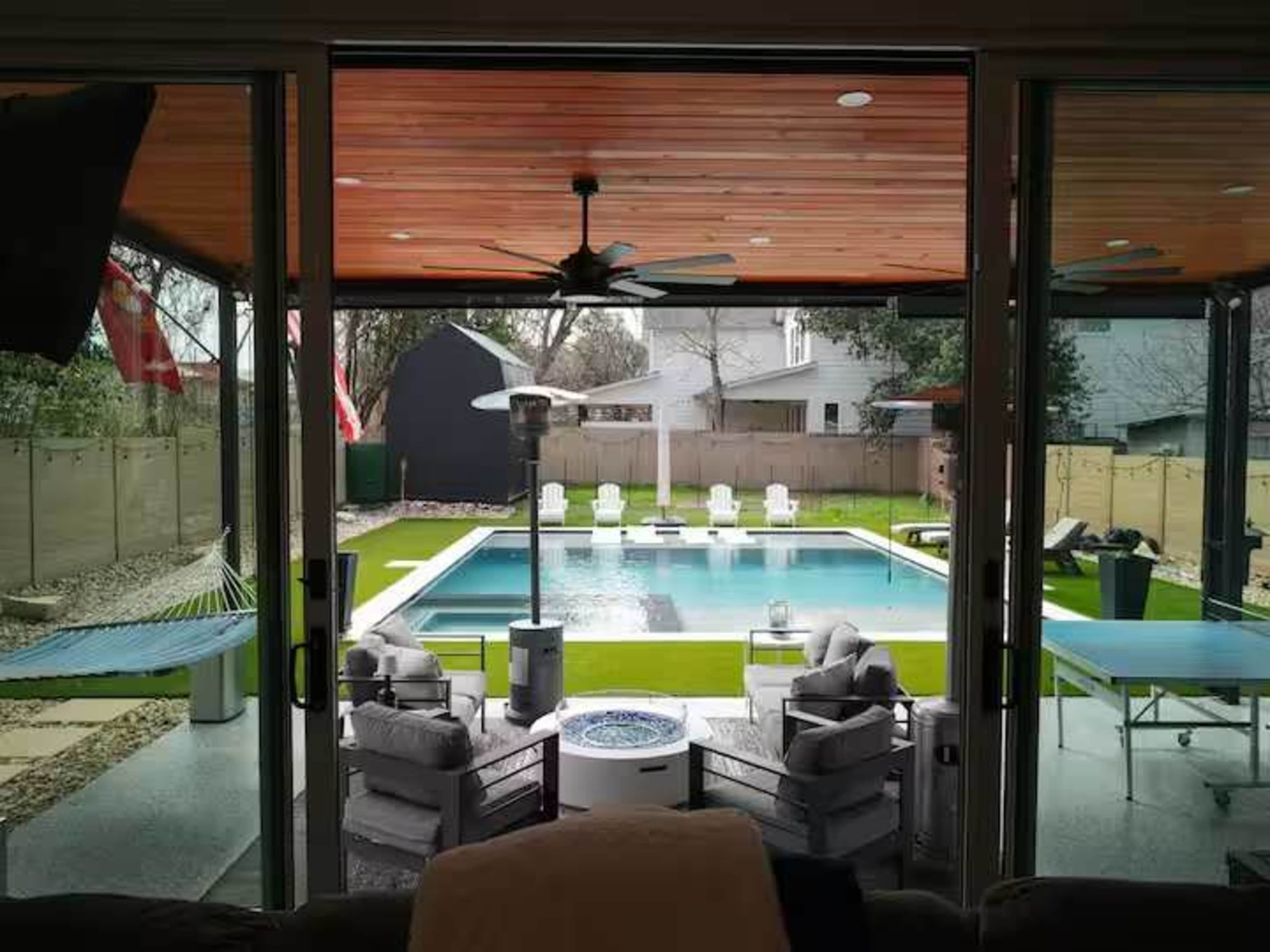 The view from the interior of a house shows a swimming pool surrounded by lounge chairs and a grassy area, with a hammock and patio heaters visible.