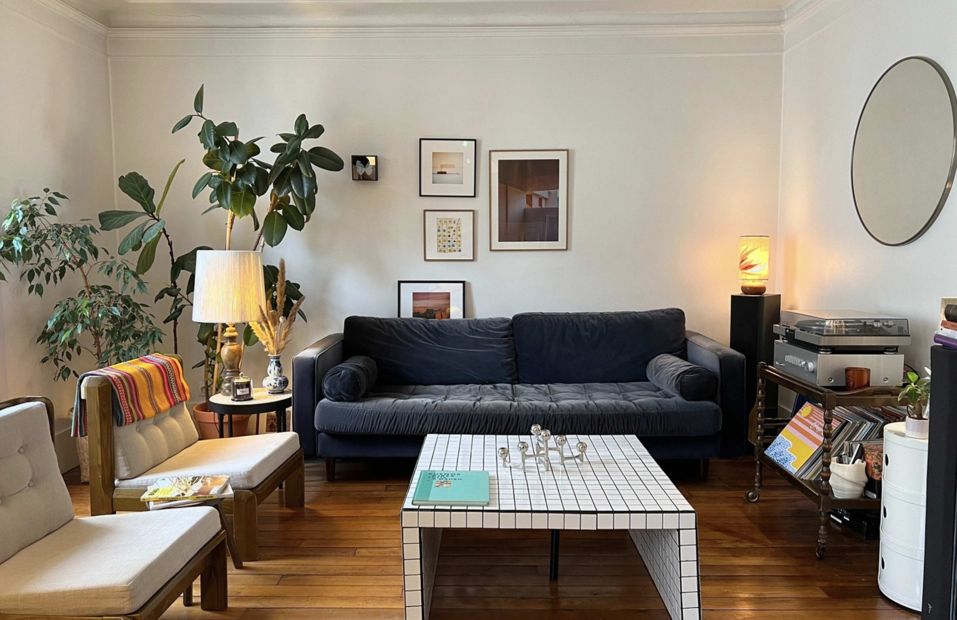 The image shows a cozy living room with a blue couch, a coffee table with a grid design, various houseplants, framed artwork on the walls, and a turntable next to a stack of records.