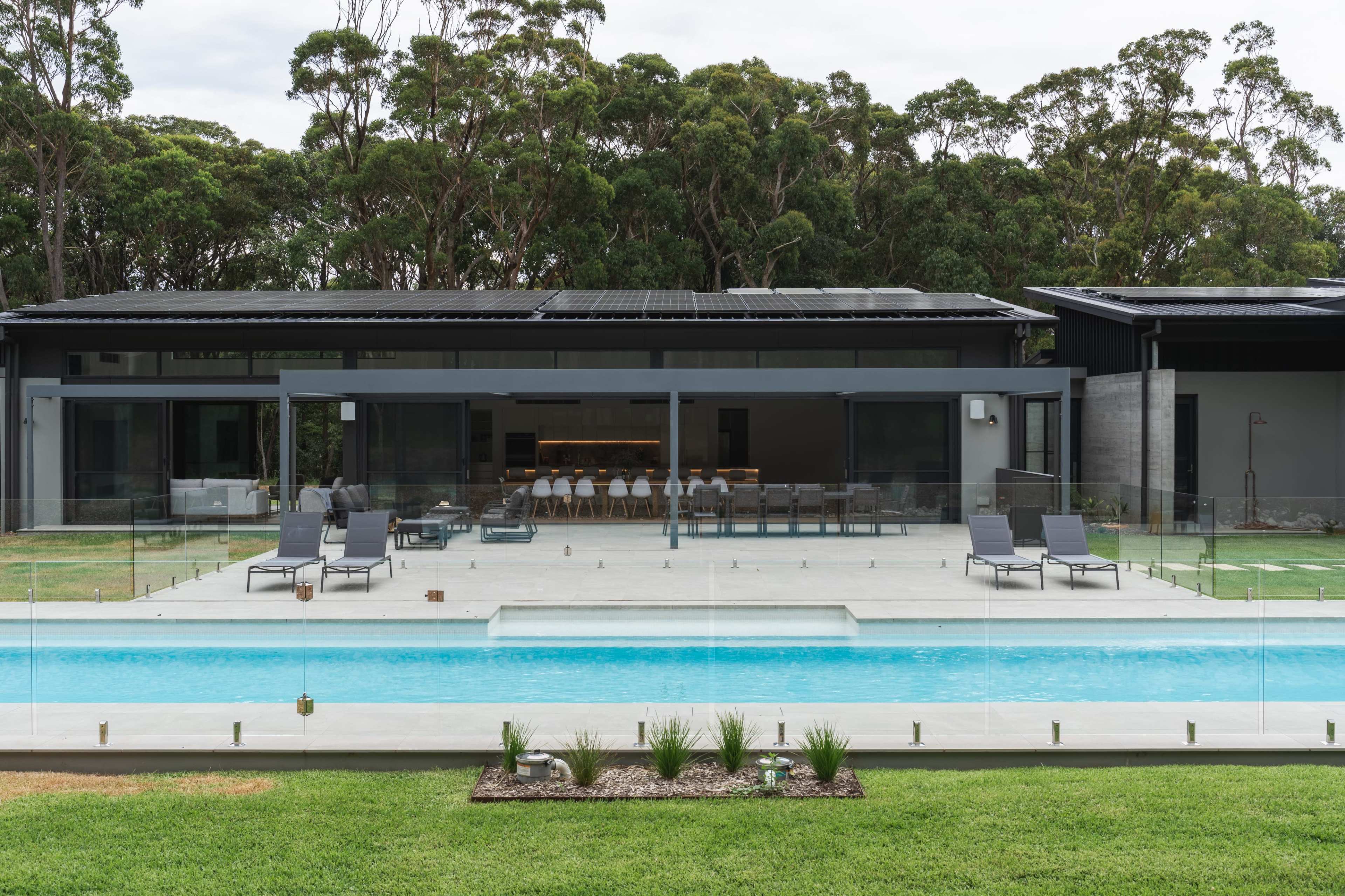 A modern home with a large glass-panelled back porch, overlooking a swimming pool surrounded by a manicured lawn and dense trees in the background.