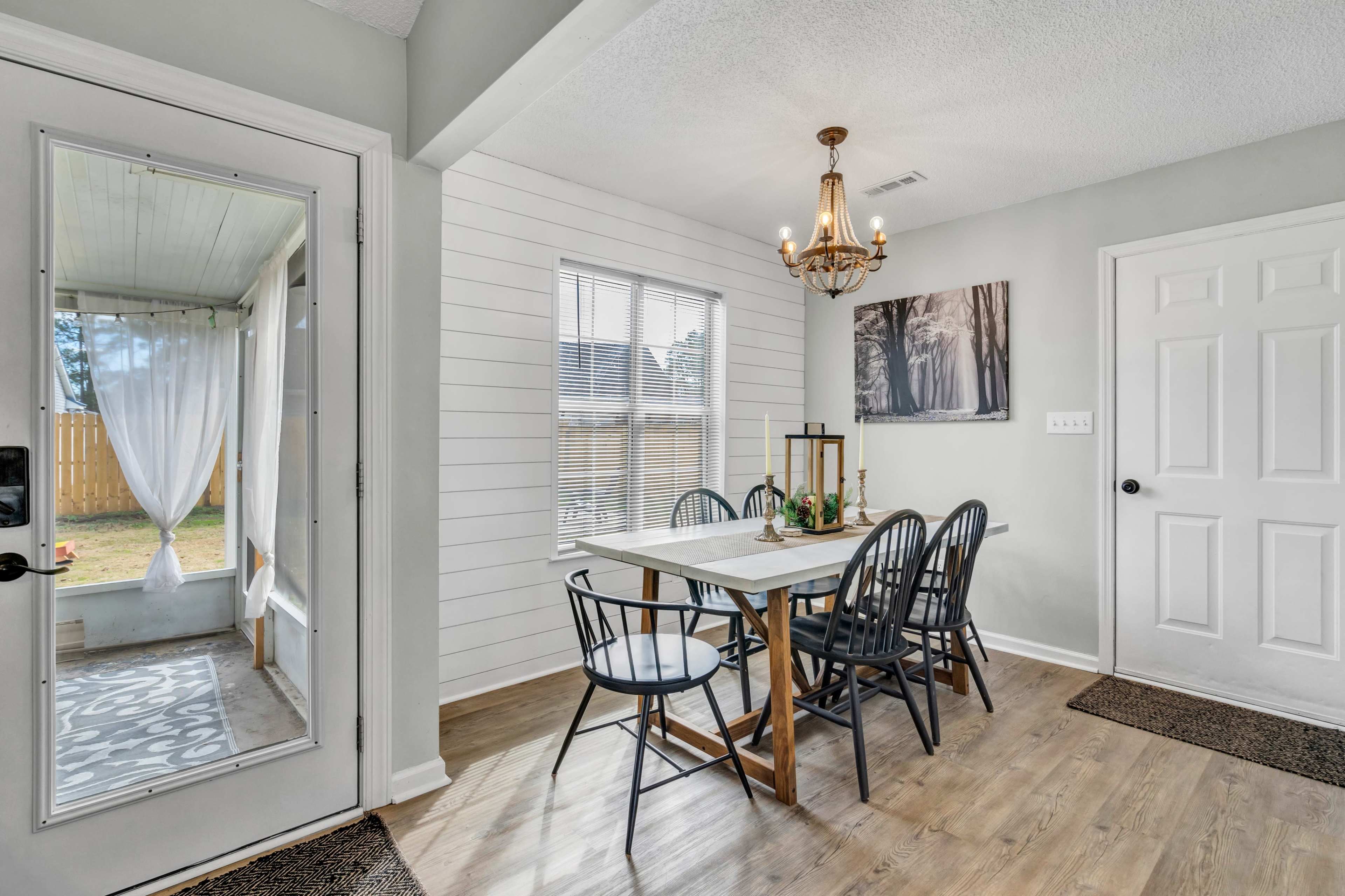 A dining area features a wooden table surrounded by black chairs, a chandelier above, and large windows letting in natural light.