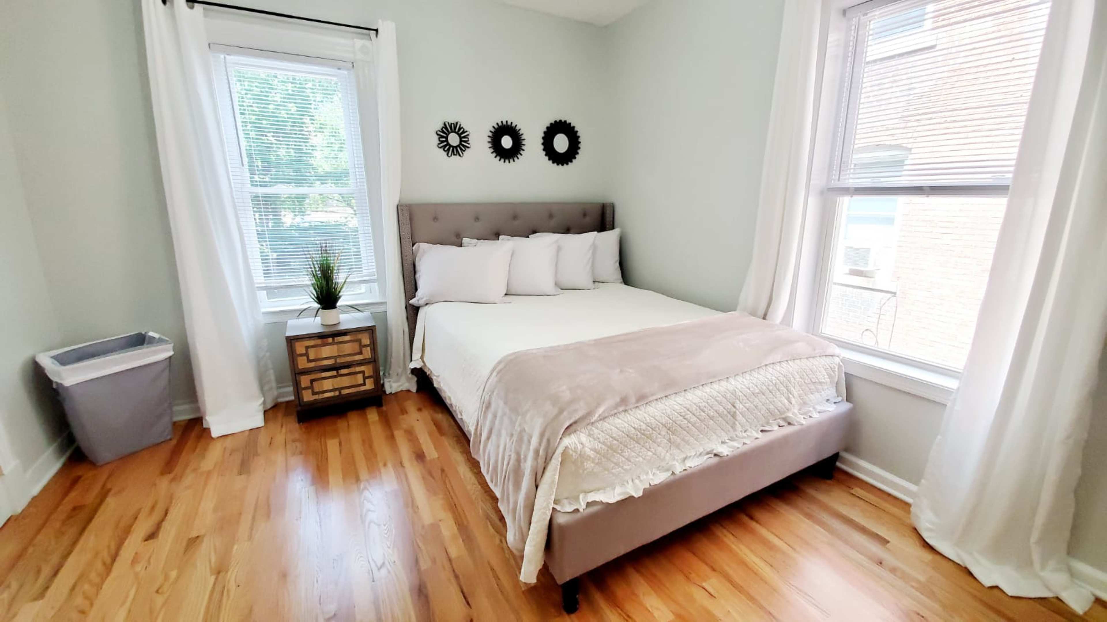 The image shows a neatly arranged bedroom featuring a bed with light bedding, a small nightstand, and large windows allowing natural light.