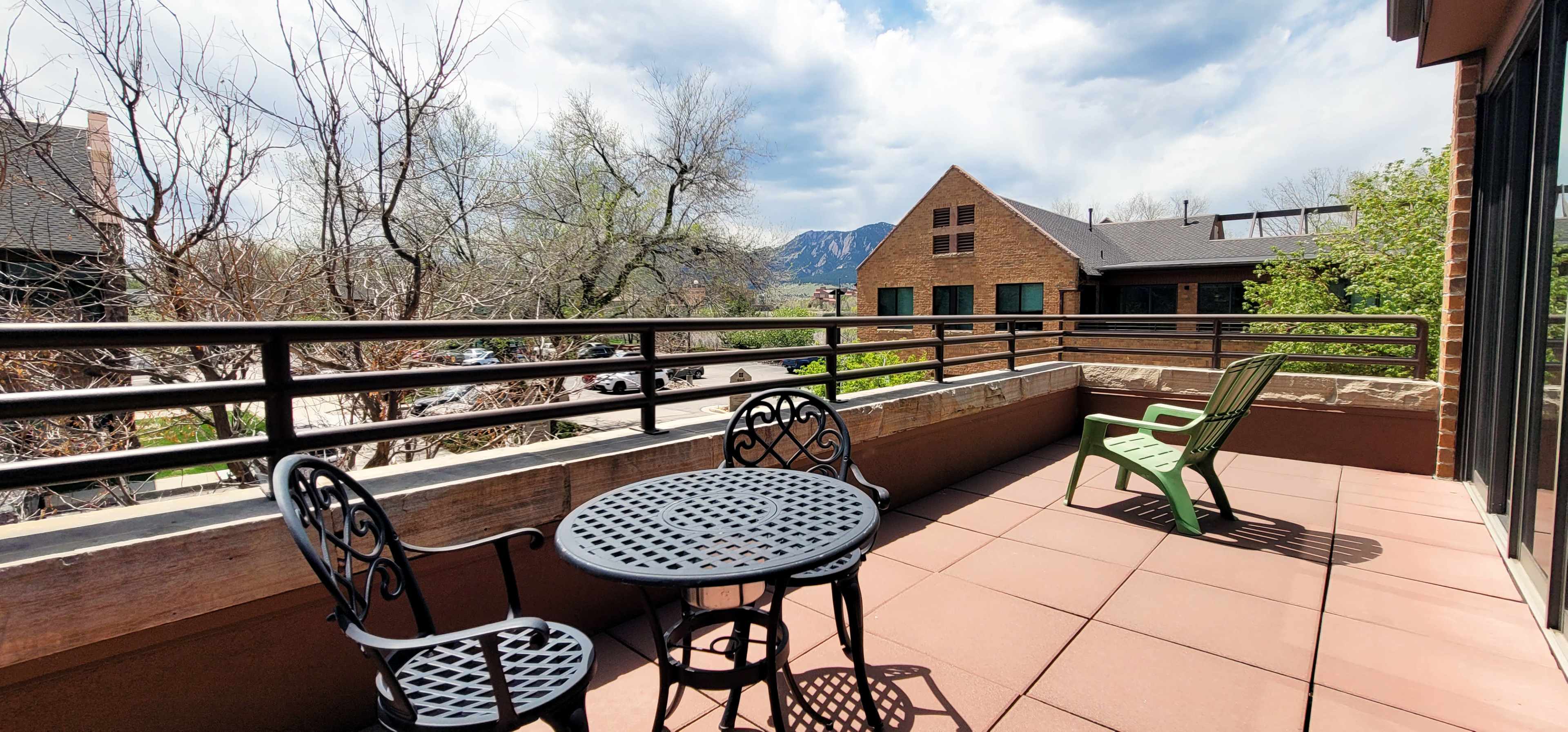 A balcony features a round table and two chairs, overlooking a street lined with trees and buildings, with mountains in the background under a partly cloudy sky.