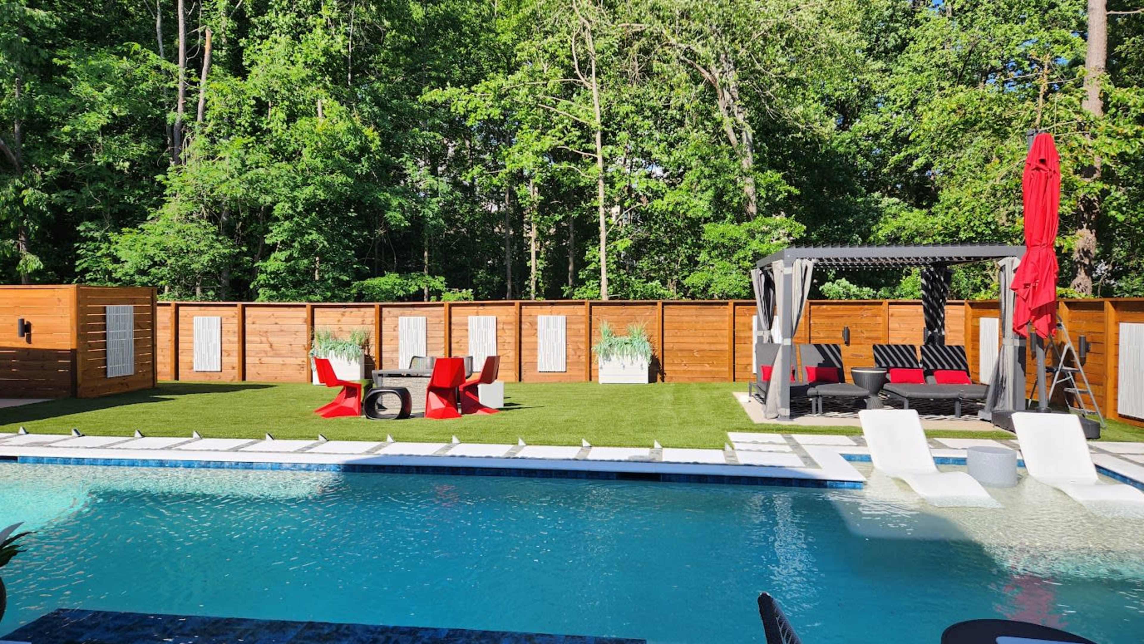 The image shows a backyard with a swimming pool, lounge chairs, a seating area with red chairs and a canopy, and wooden fencing surrounded by lush greenery.