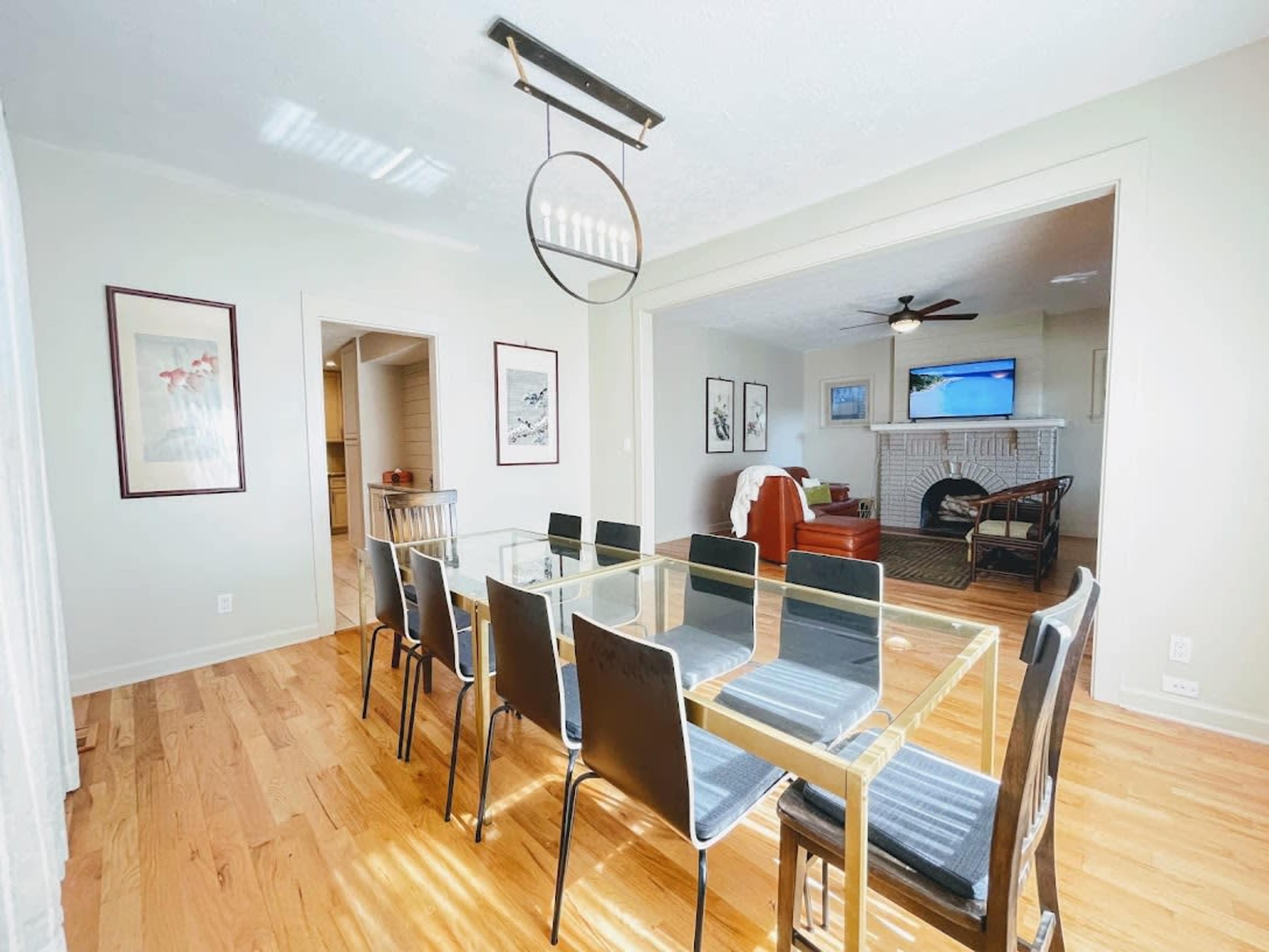 A dining area features a glass table surrounded by black chairs, with a view of a living room that includes a fireplace and a television.