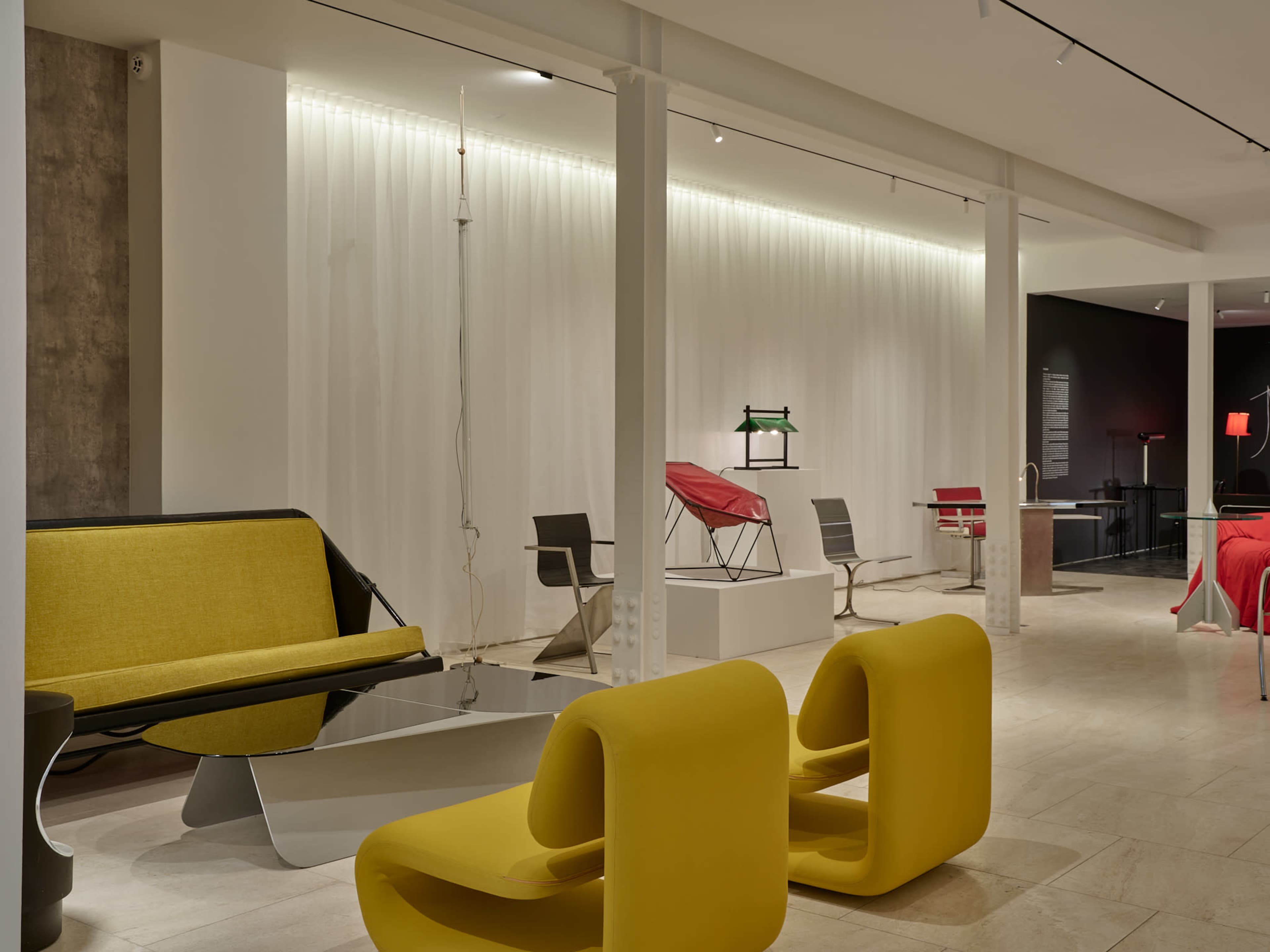 The image shows a modern interior space featuring minimalist furniture, including a yellow sofa, stylized chairs, and a unique coffee table, with soft lighting illuminating the area.