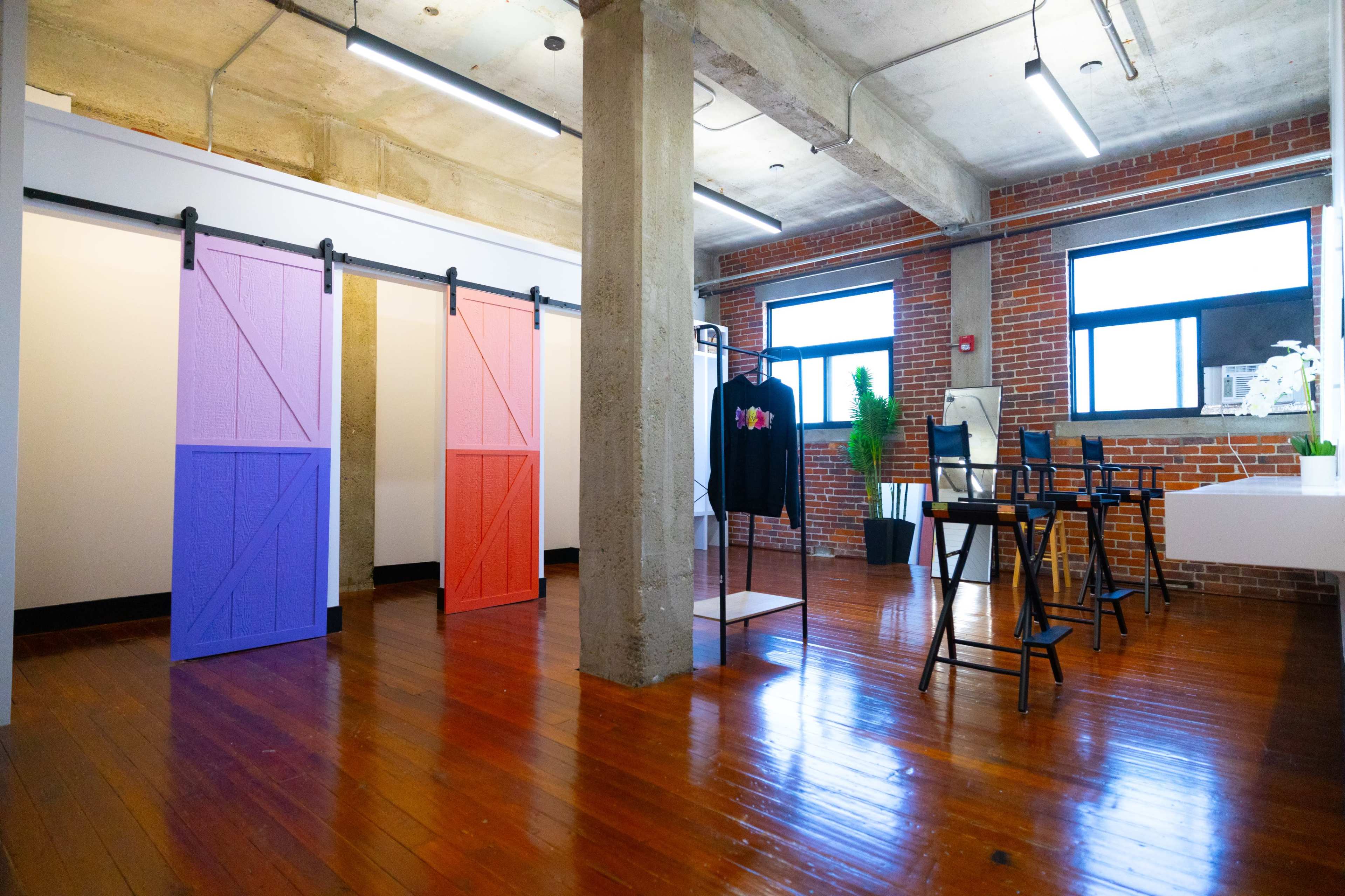 An industrial-style room with exposed brick walls, wooden floors, a table and chairs, and two sliding barn doors, one pink and one purple, separating different areas.