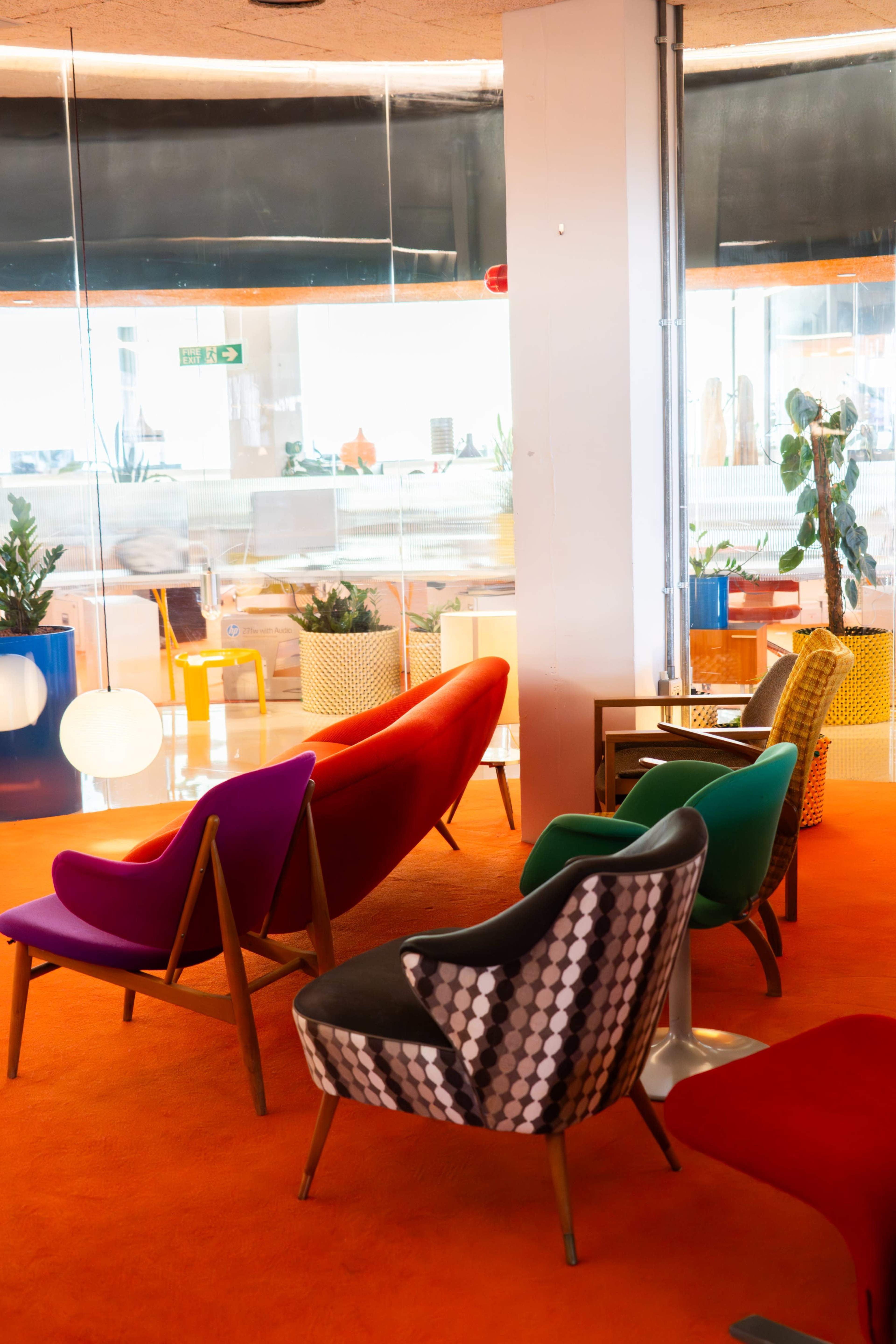 The image features a colorful lounge area with various modern chairs, a table, and plants, set against a backdrop of large windows.