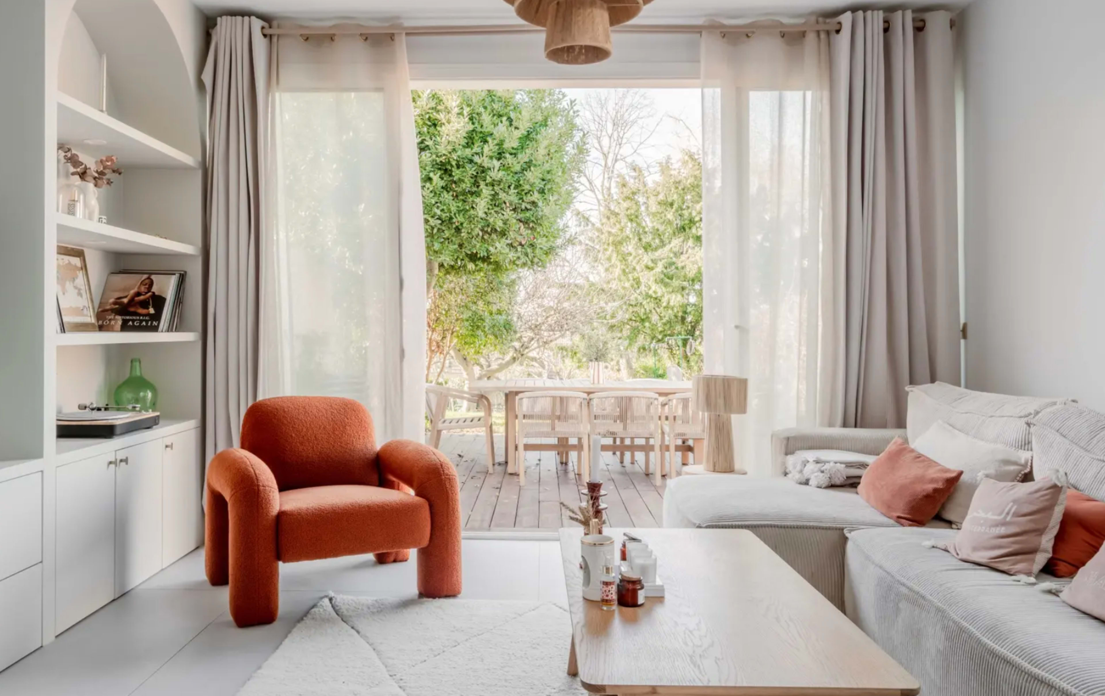 A cozy living room with a light-colored sofa, an orange chair, a wooden coffee table, and large windows that open to a patio with greenery.
