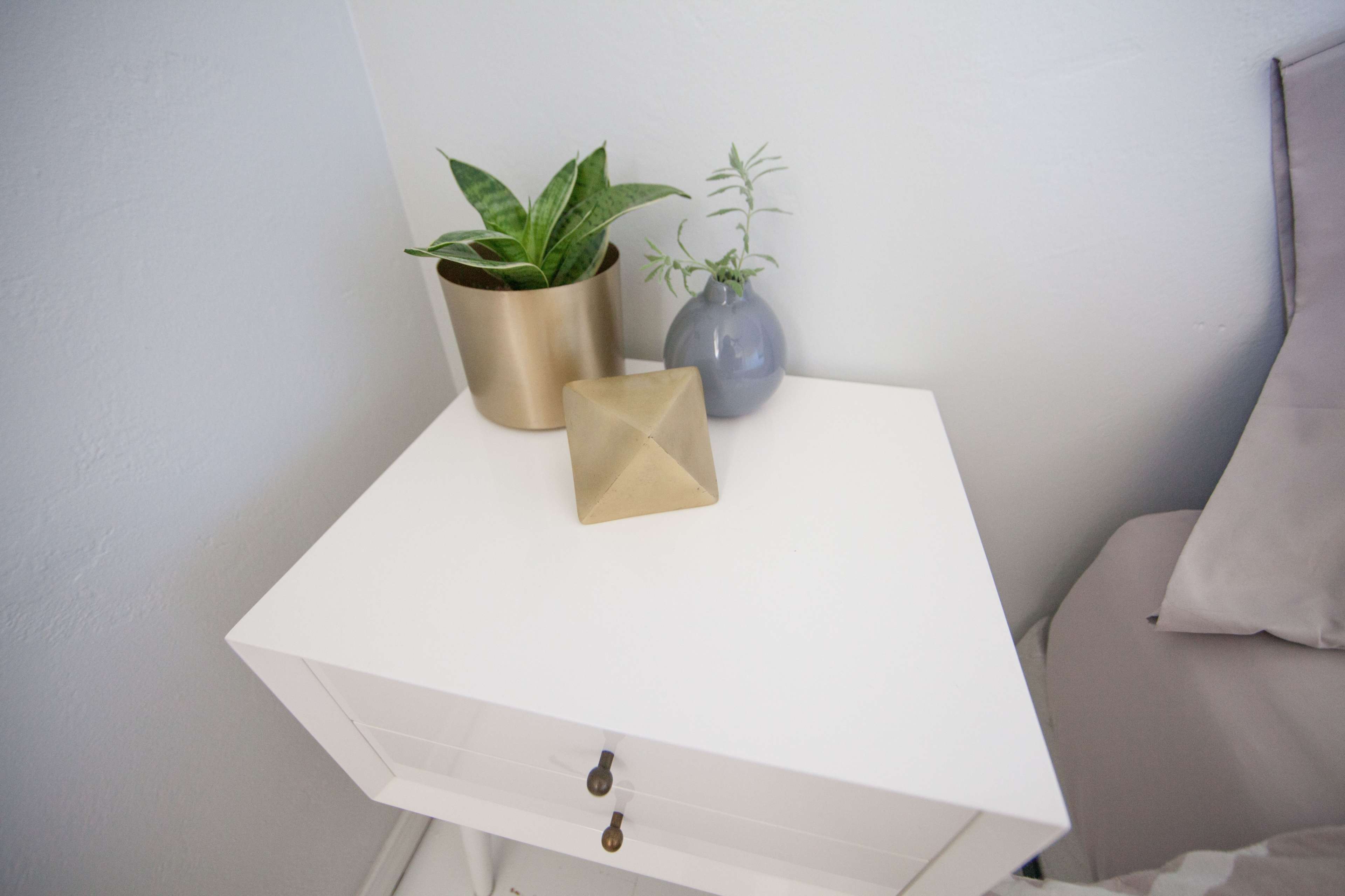 A white bedside table holds a gold and gray decorative object alongside two potted plants.
