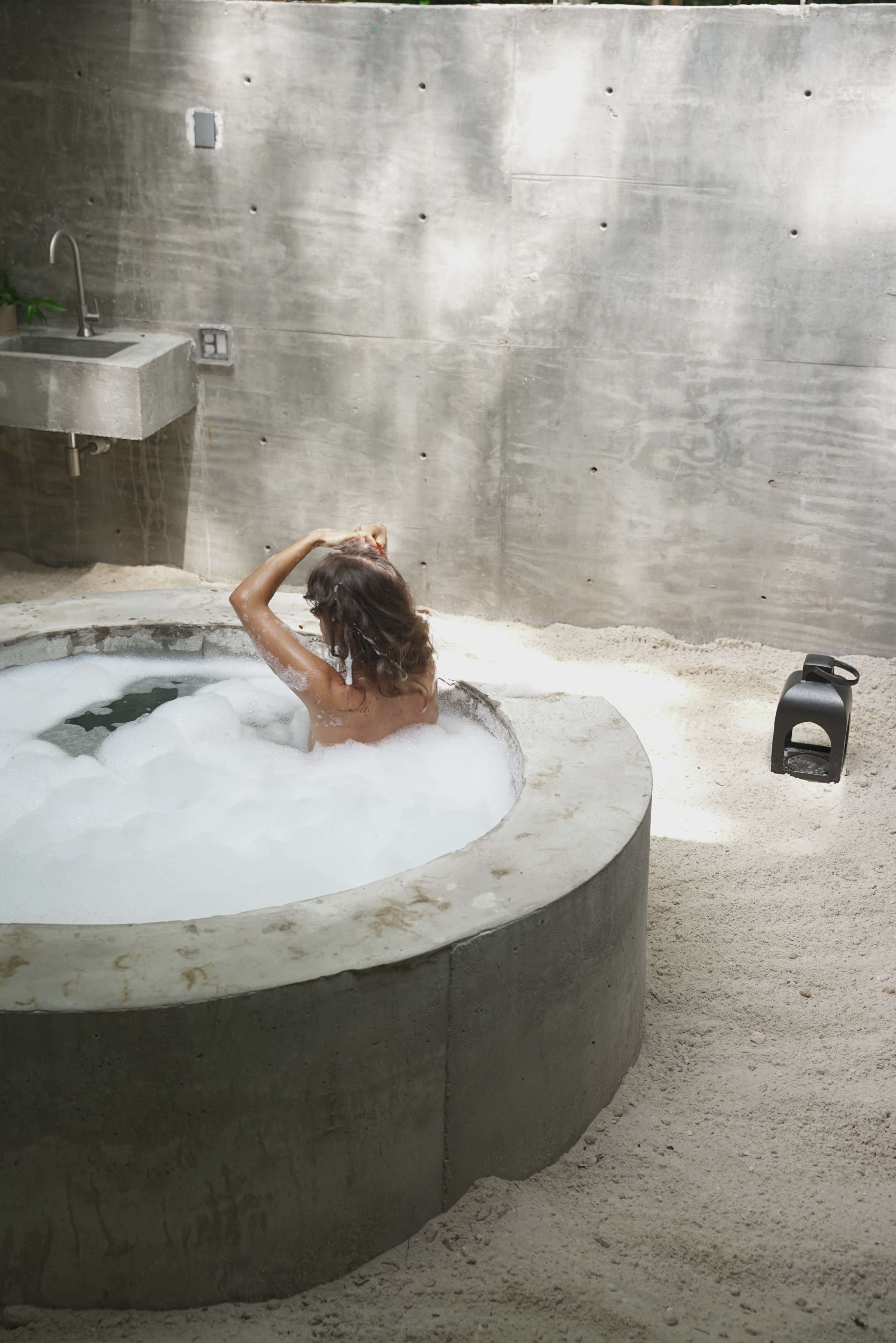 A woman with curly hair sits in a circular freestanding bathtub filled with bubbles in a minimalist bathroom with concrete walls and sandy flooring.
