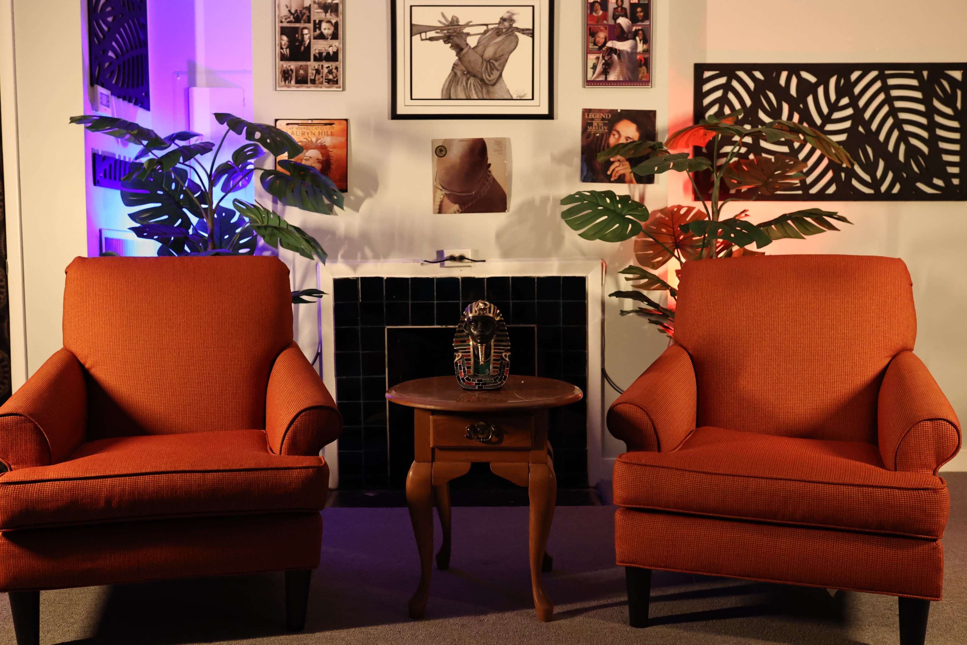 Two orange armchairs facing a small wooden table in a room decorated with various framed photos and plants, with purple and blue lighting.