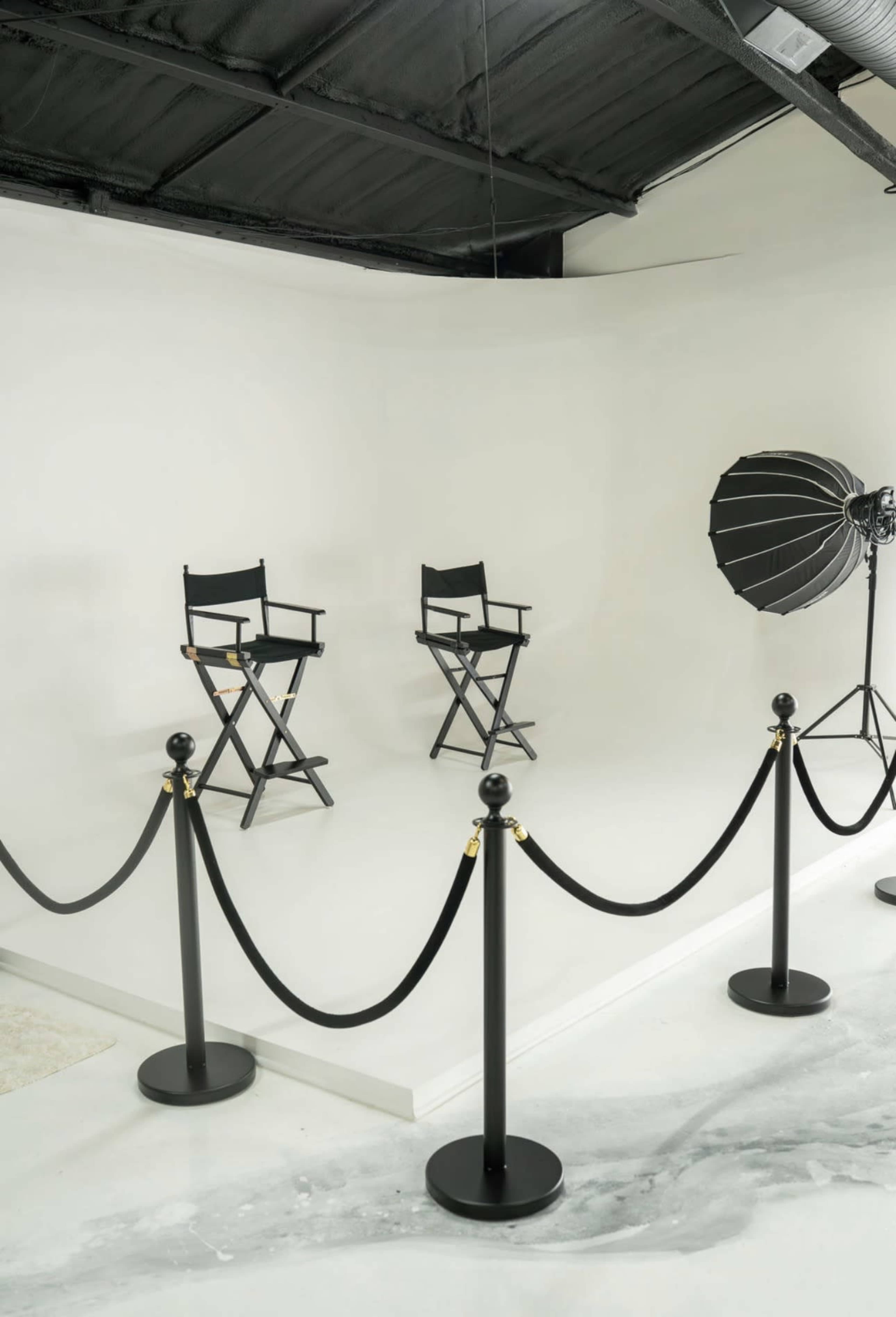 The image shows a minimalist studio setup featuring two director's chairs, a large softbox light, and a rope barrier enclosing the area.