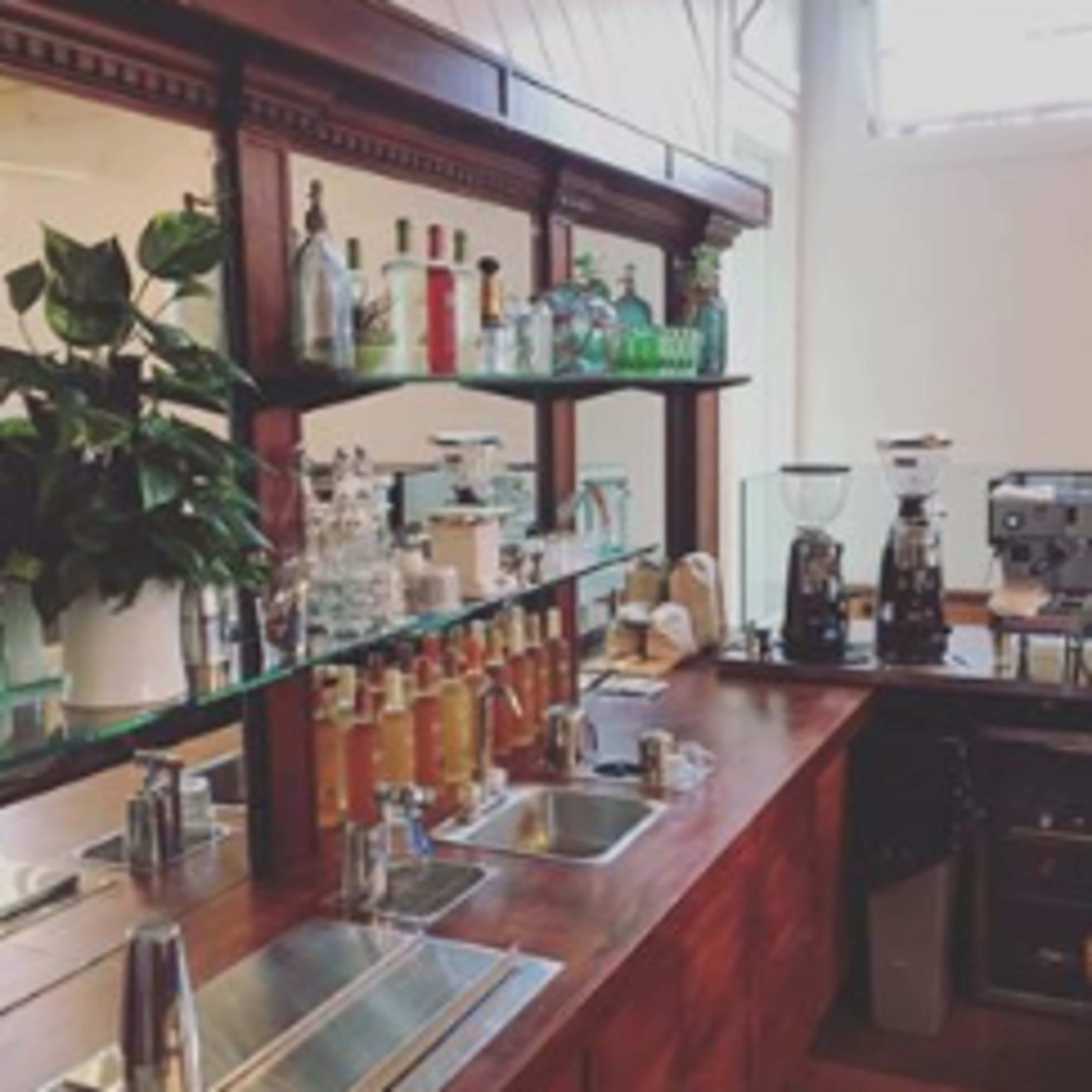 The image shows a wooden bar with a selection of glass bottles, a coffee grinder, and a plant in a white pot.
