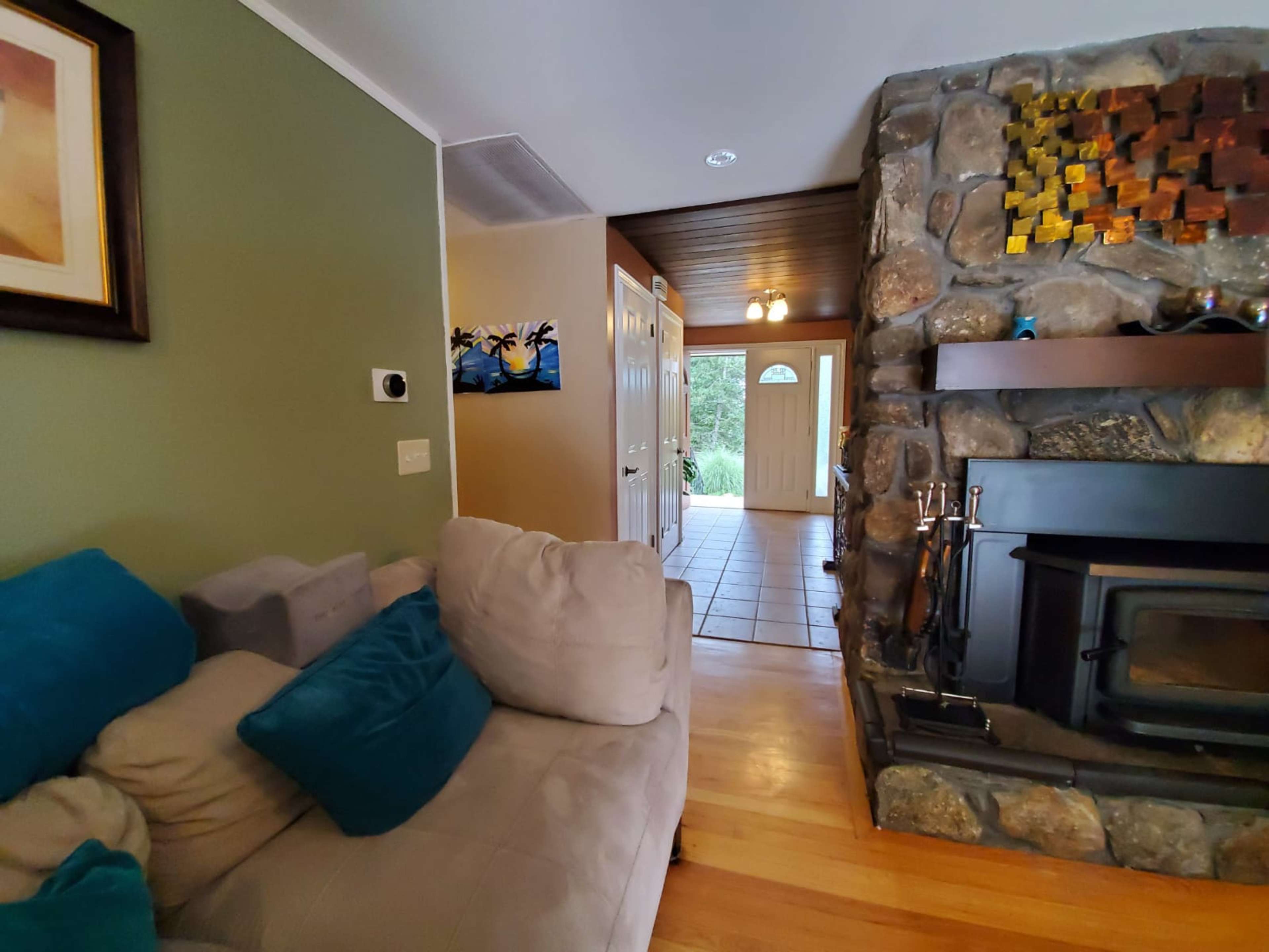 A cozy living room with a stone fireplace and a light-colored couch adorned with blue pillows, leading to a door that opens to an outdoor area.