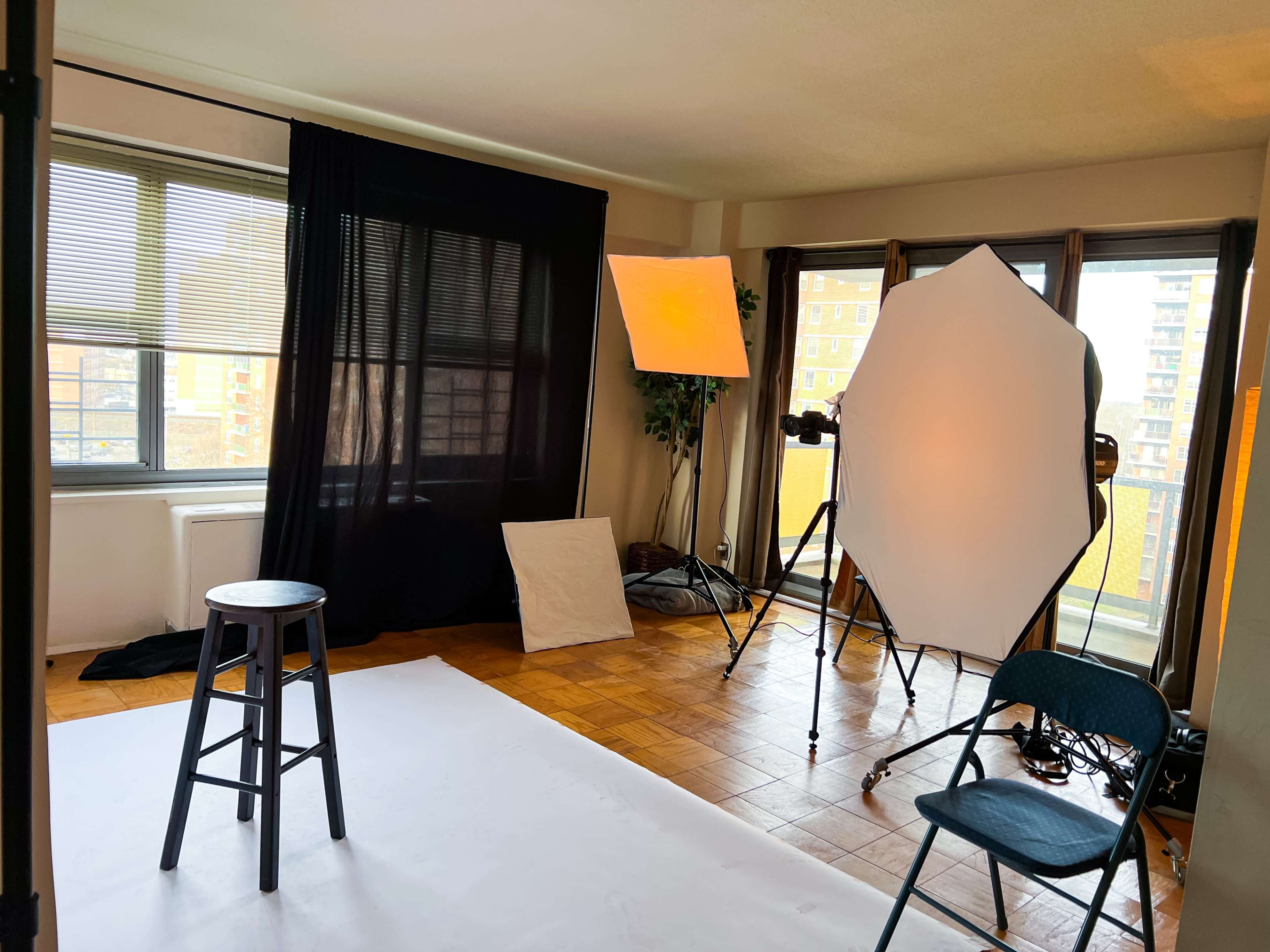 A well-lit photography studio setup featuring a black backdrop, a stool, and softbox lights arranged next to a large window.