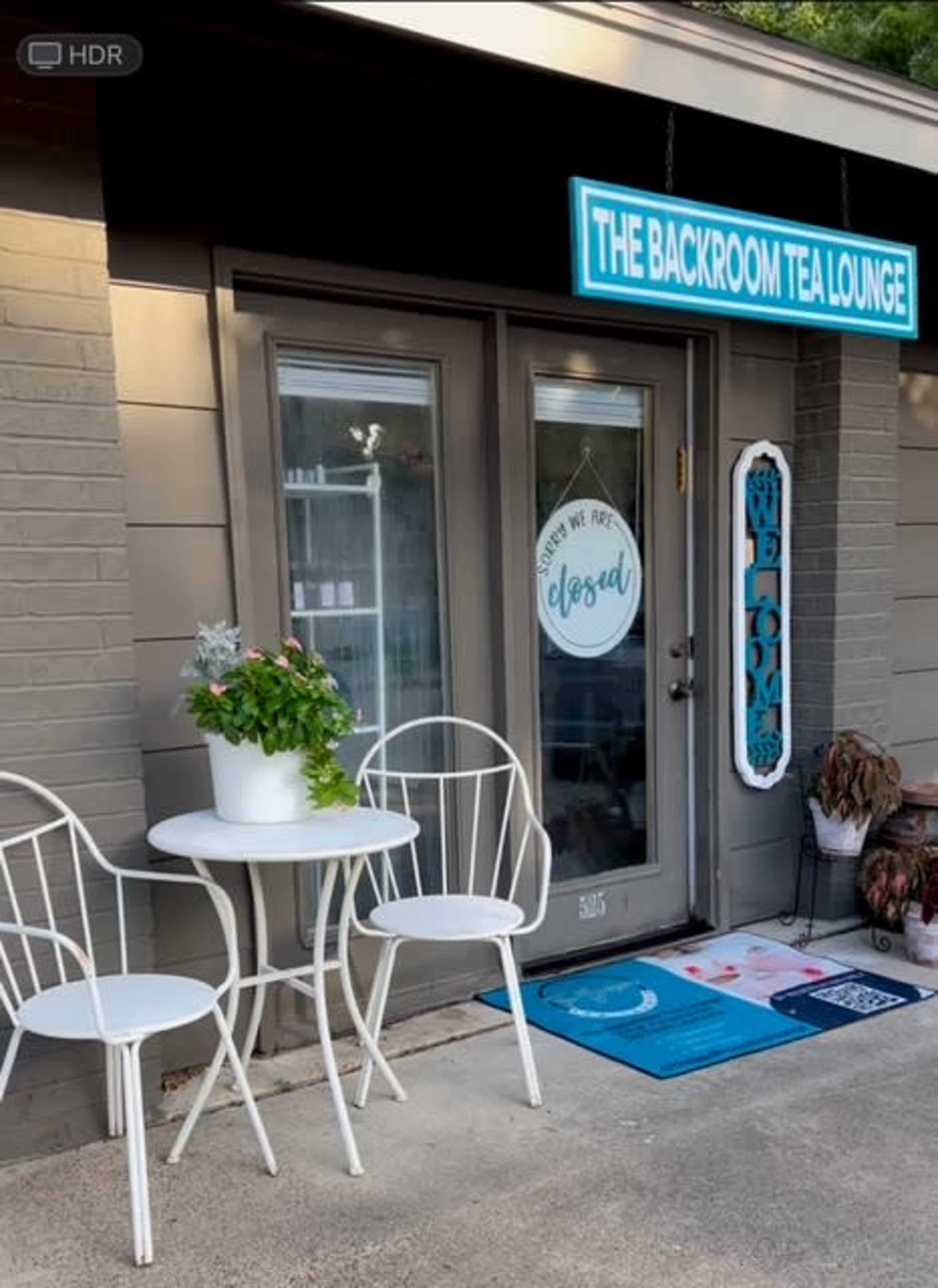 The image shows the entrance of a tea lounge named "The Backroom Tea Lounge," featuring a small outdoor seating area with two white chairs and a table.