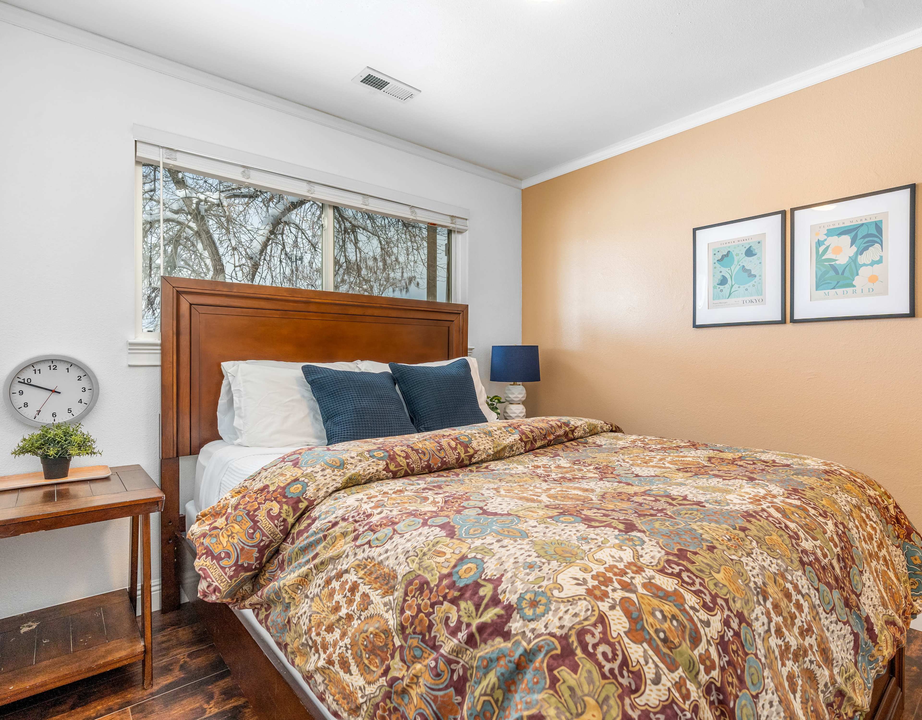 A cozy bedroom features a wooden bed with a colorful patterned comforter, a bedside table, a lamp, and framed artwork on the wall.