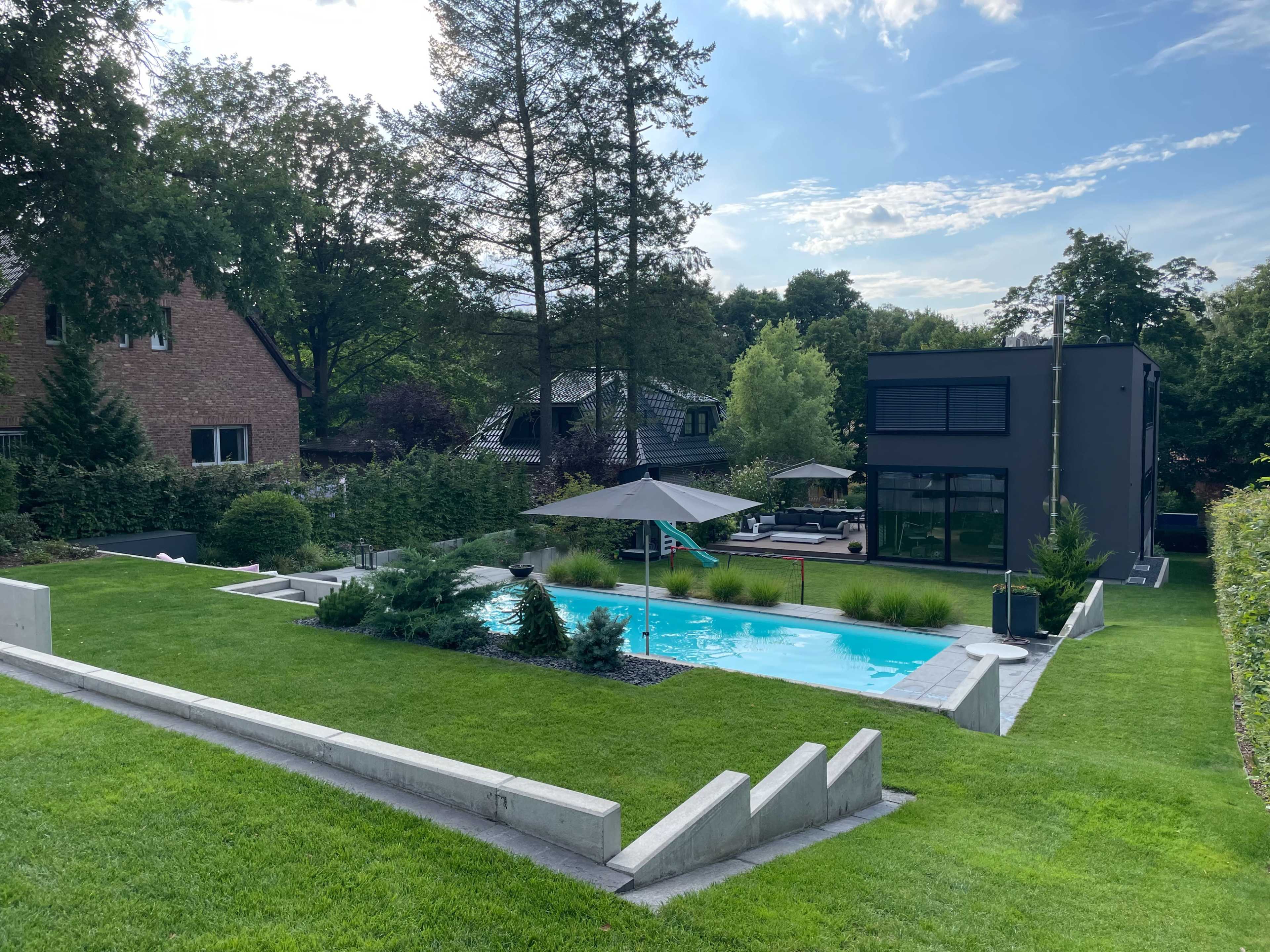 A landscaped backyard with a swimming pool, surrounded by green grass, trees, and a modern two-story building in the background.