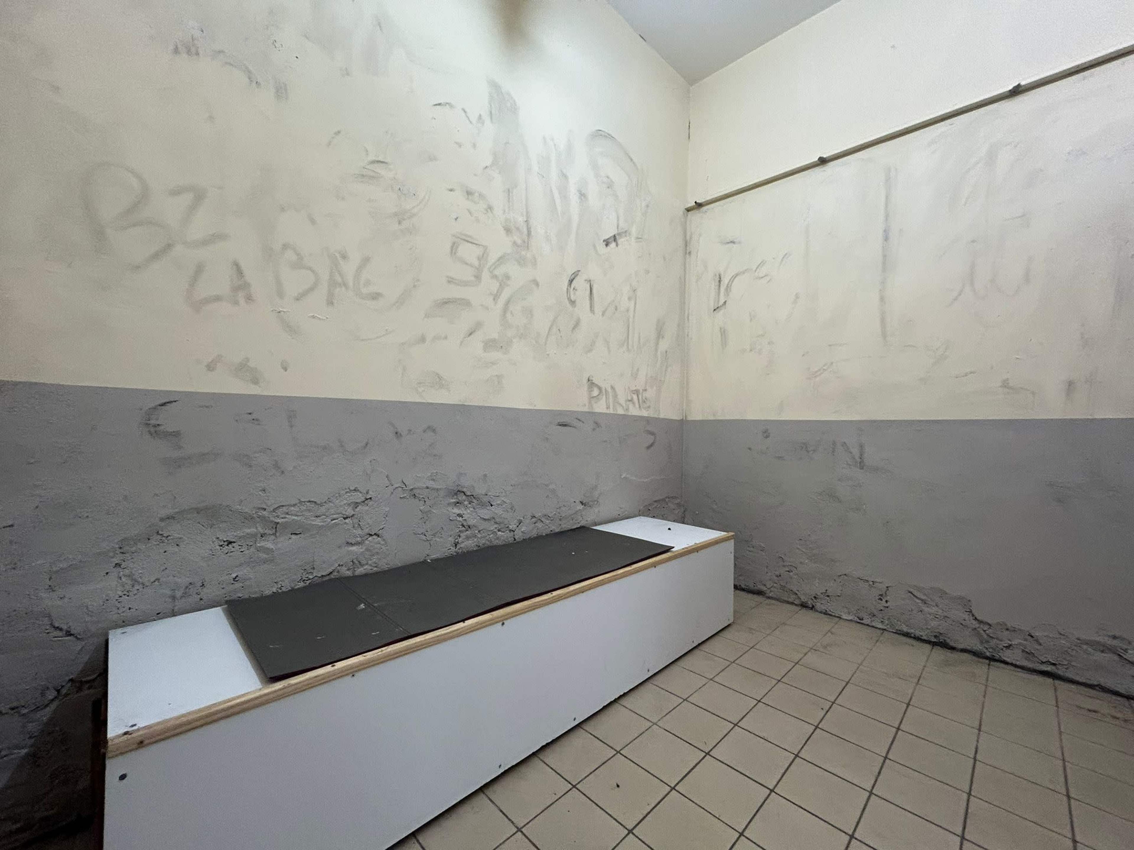 An empty, small room with a raised platform along one wall and faint graffiti on the walls.