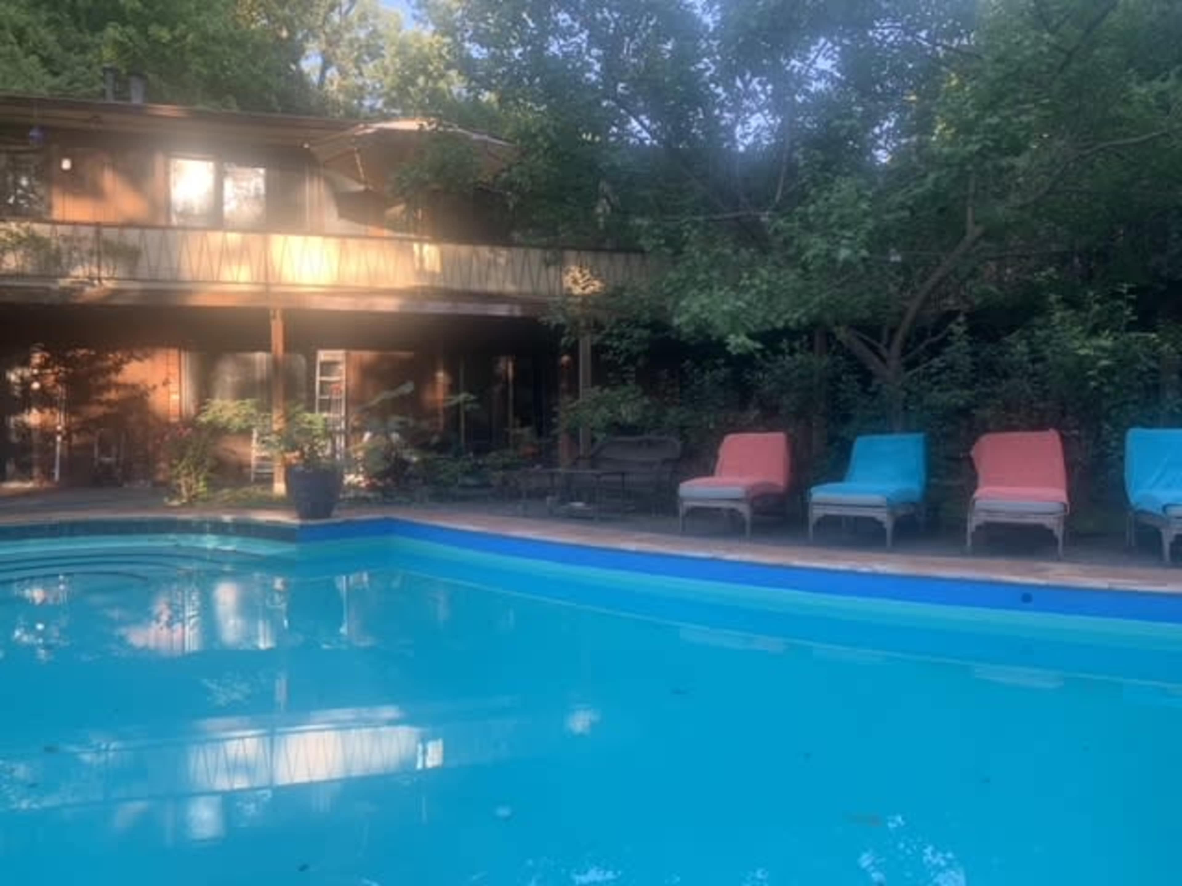 A swimming pool with clear blue water reflects the surrounding trees and a wooden deck with lounge chairs in the background.