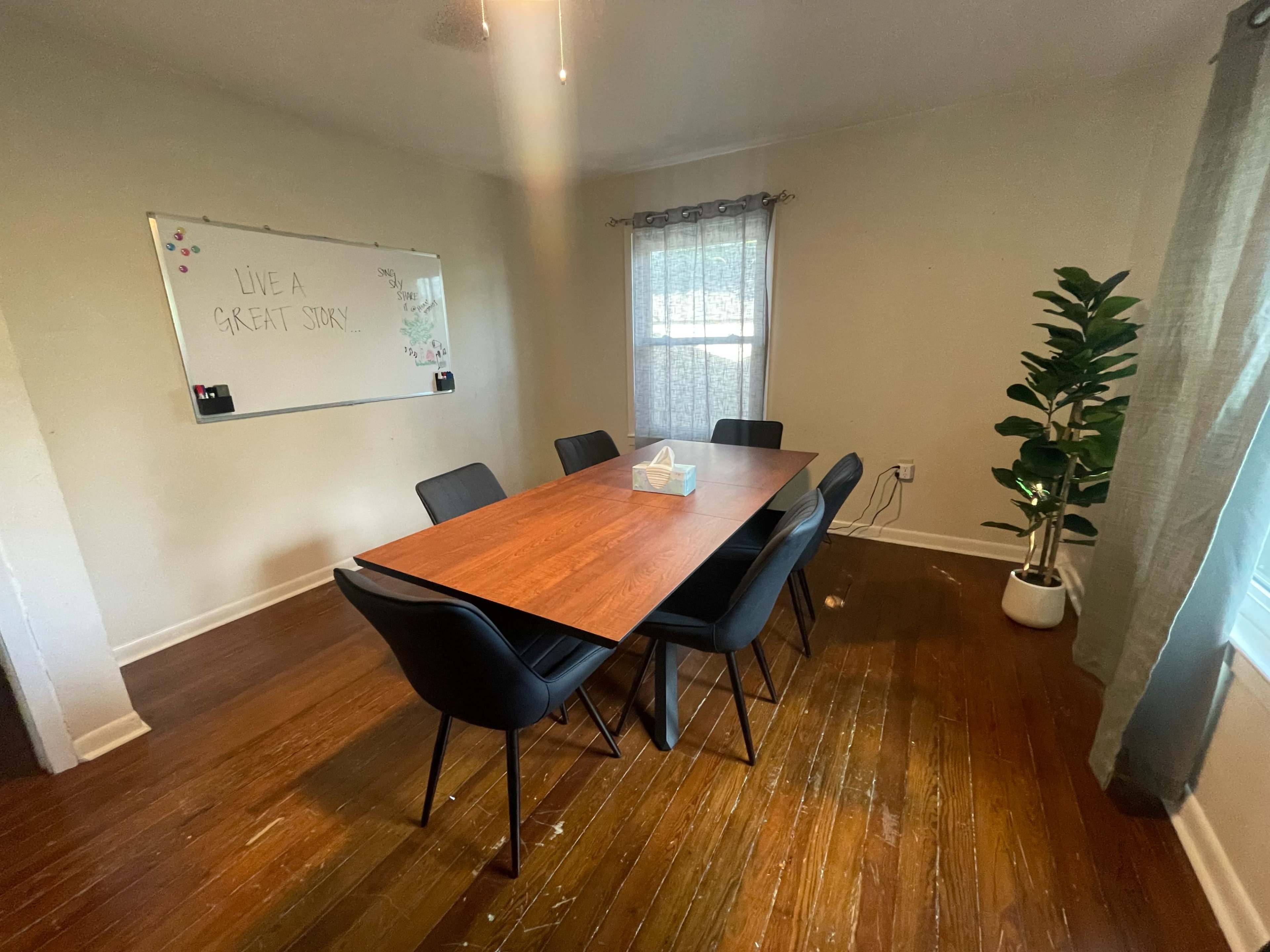 A dining room features a wooden table surrounded by black chairs, with a whiteboard on the wall and a potted plant in the corner.
