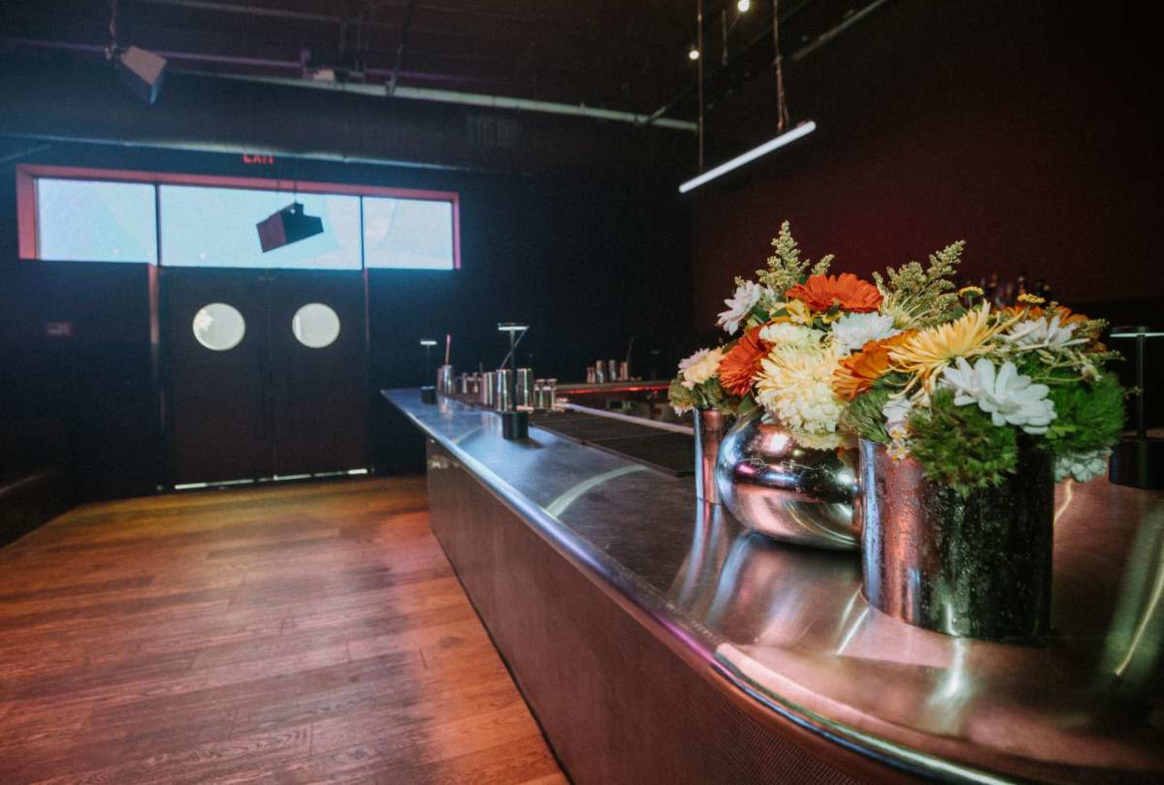 A polished bar counter with vases of flowers is situated near double doors in a dimly lit room.