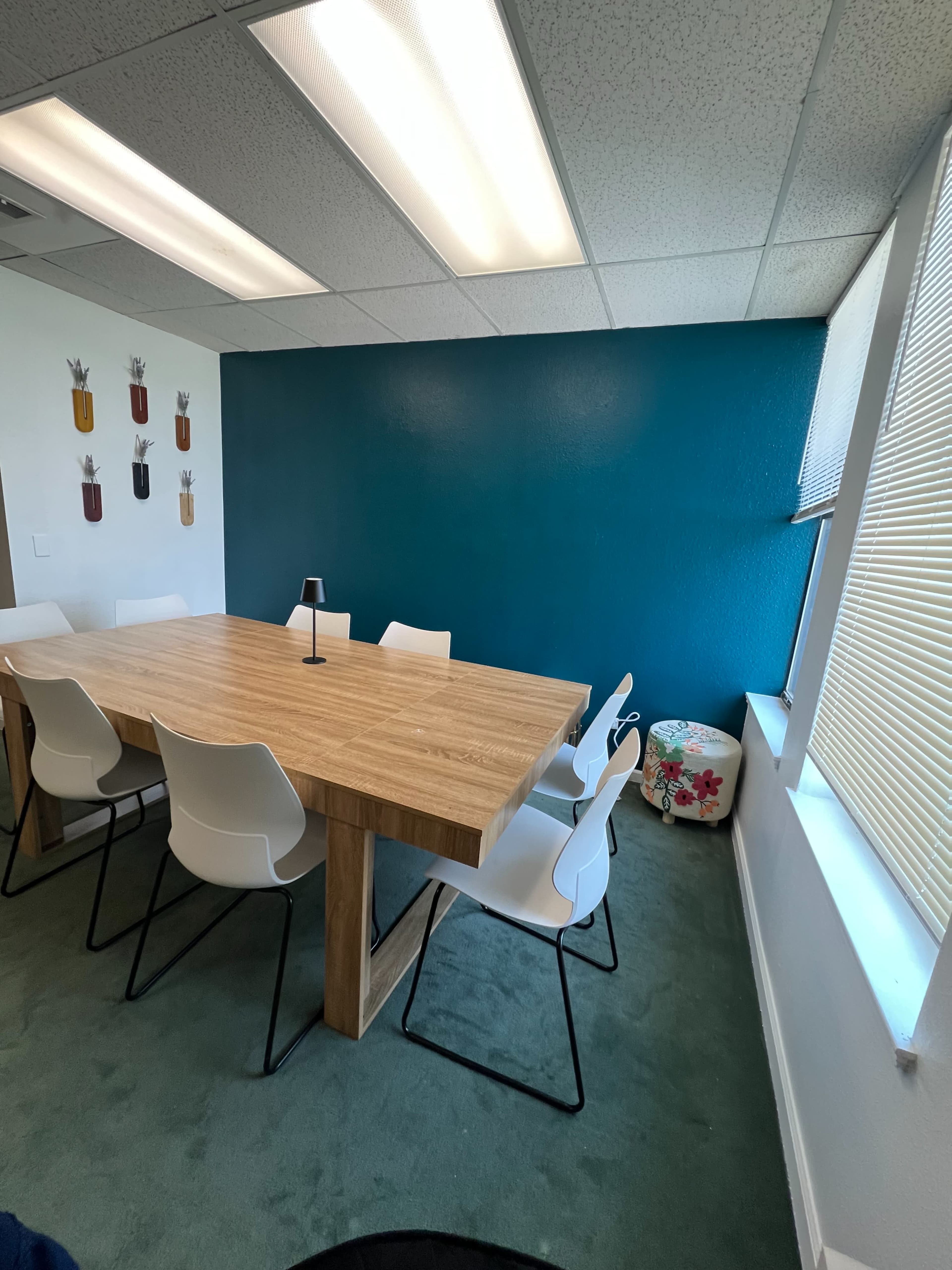 The image shows a conference room with a wooden table surrounded by white chairs, a teal wall, and a light-colored carpet.
