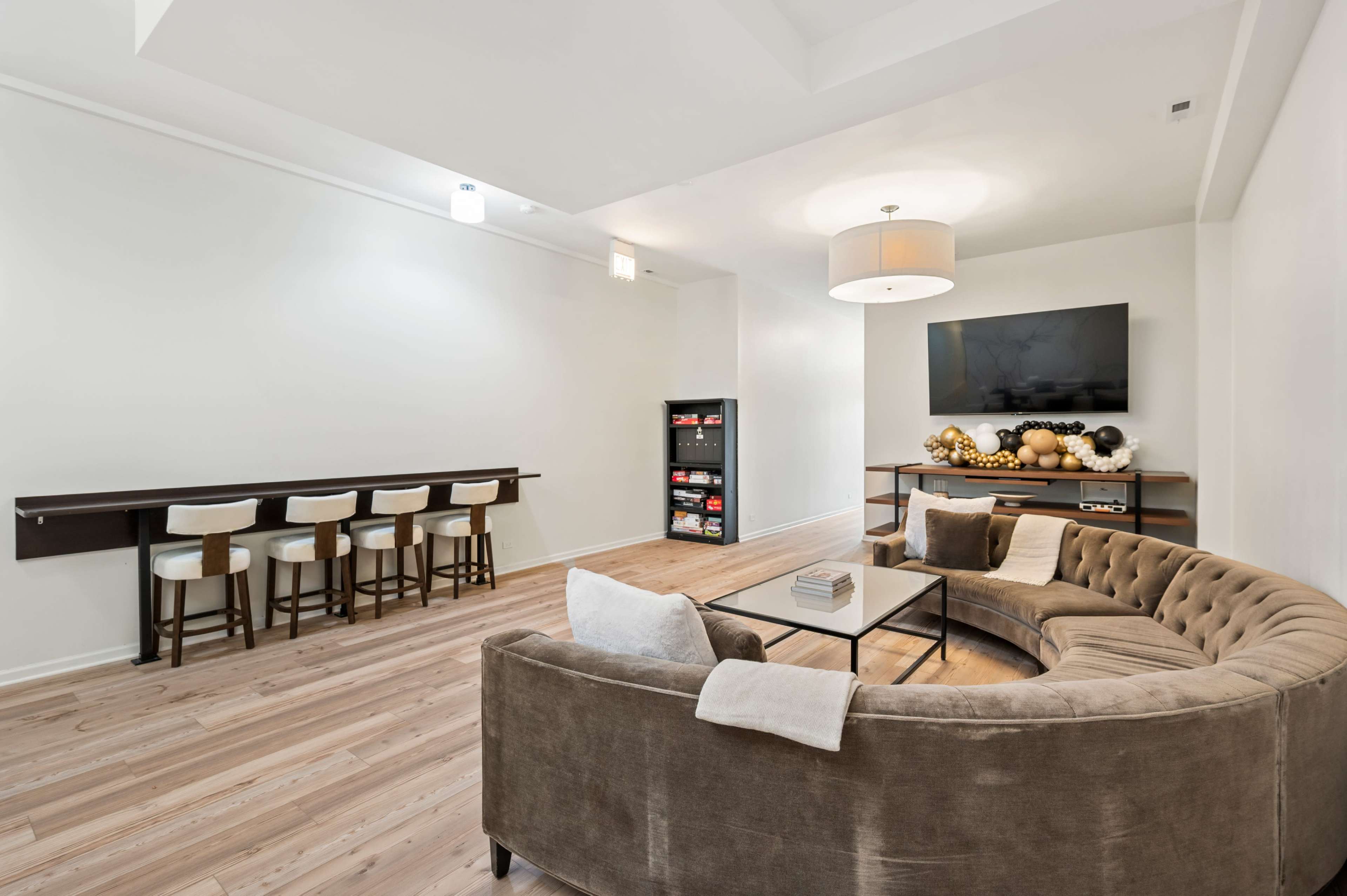 The image shows a modern living space with a curved sofa, a coffee table in the center, a wall-mounted TV, a long bar-style table with four stools, and a shelf displaying decorative items.