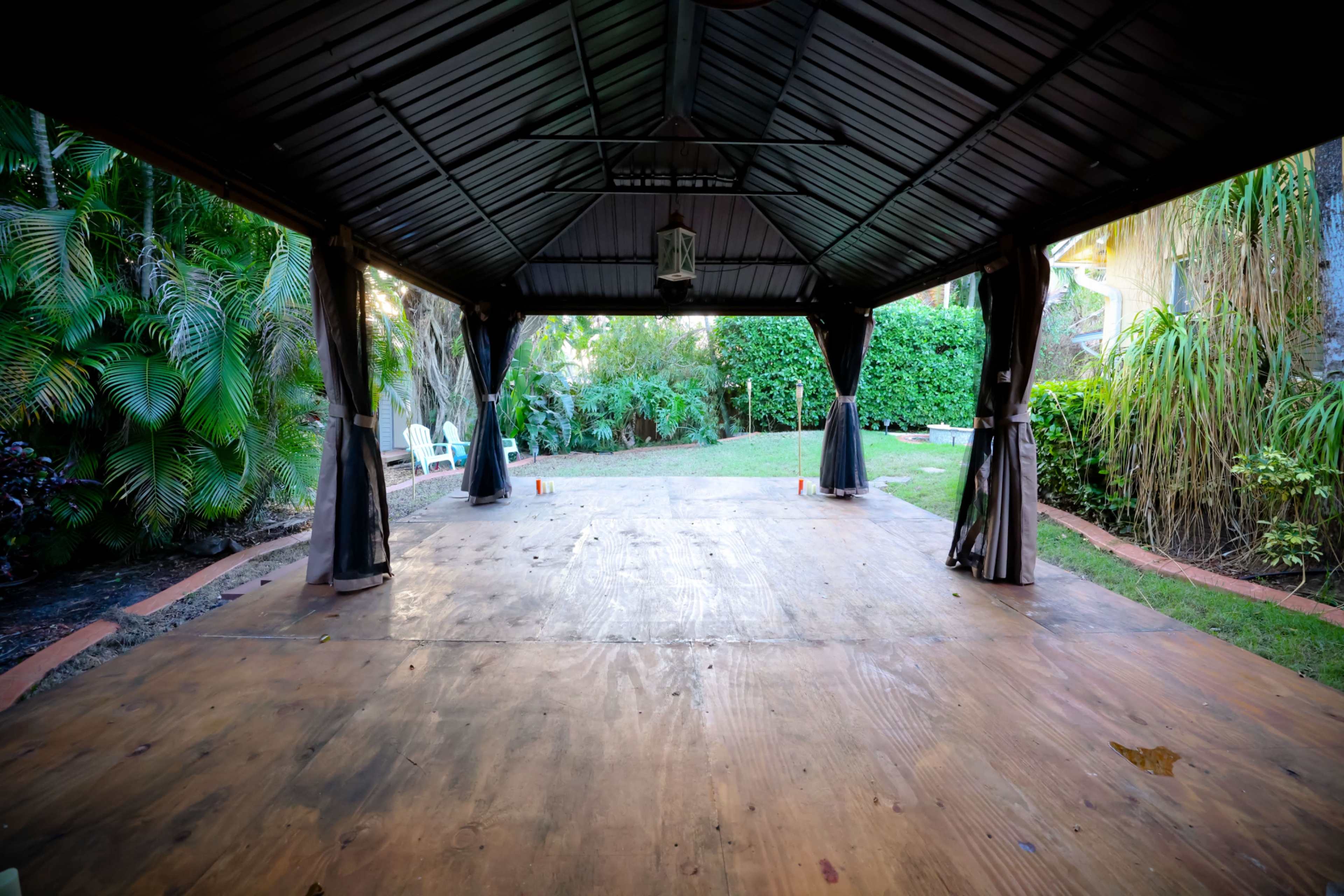 An open, covered gazebo with a wooden floor overlooks a grassy backyard surrounded by lush greenery.