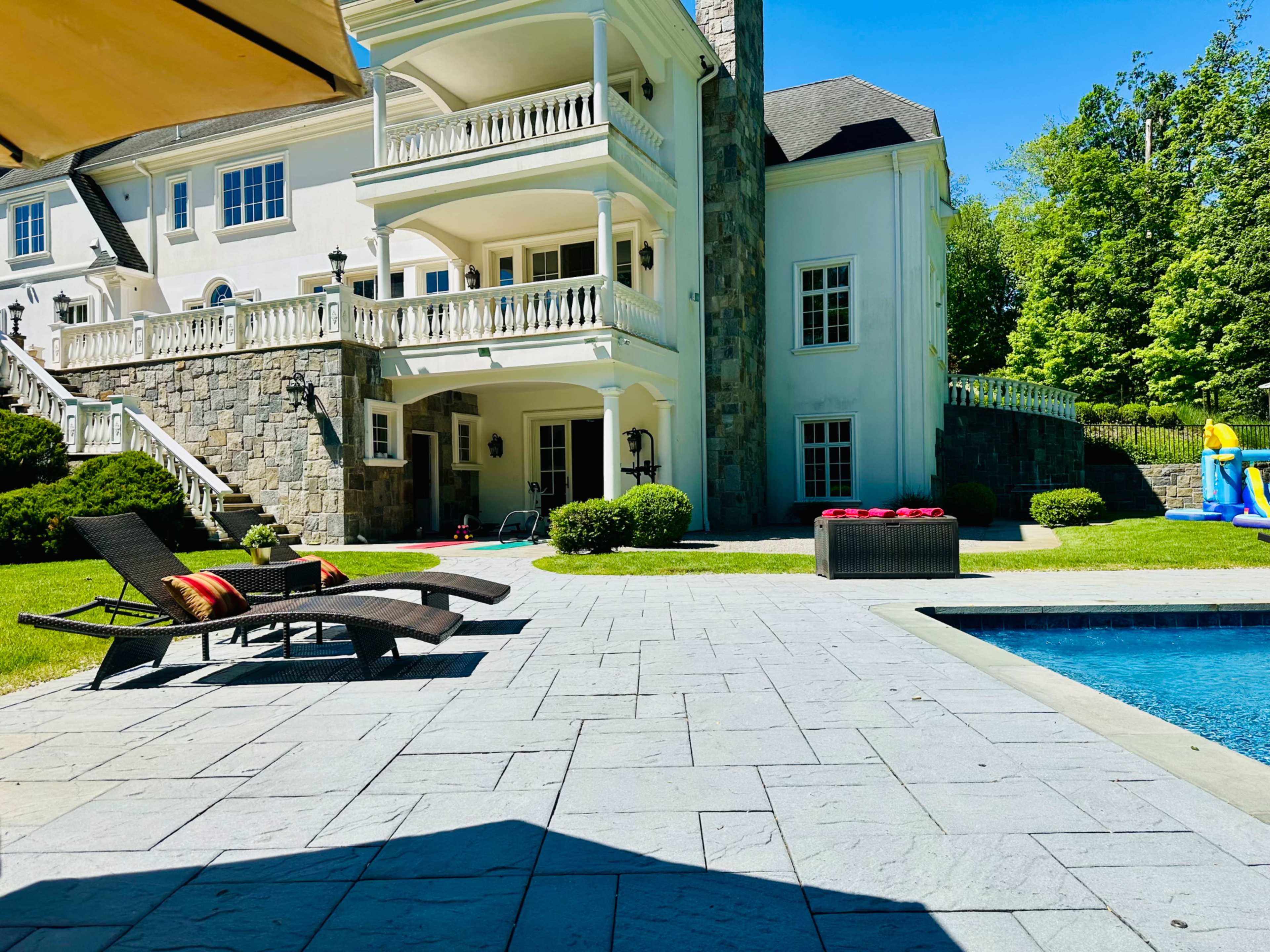 The image shows a spacious backyard with a pool, sun loungers, and a large stone house surrounded by greenery.