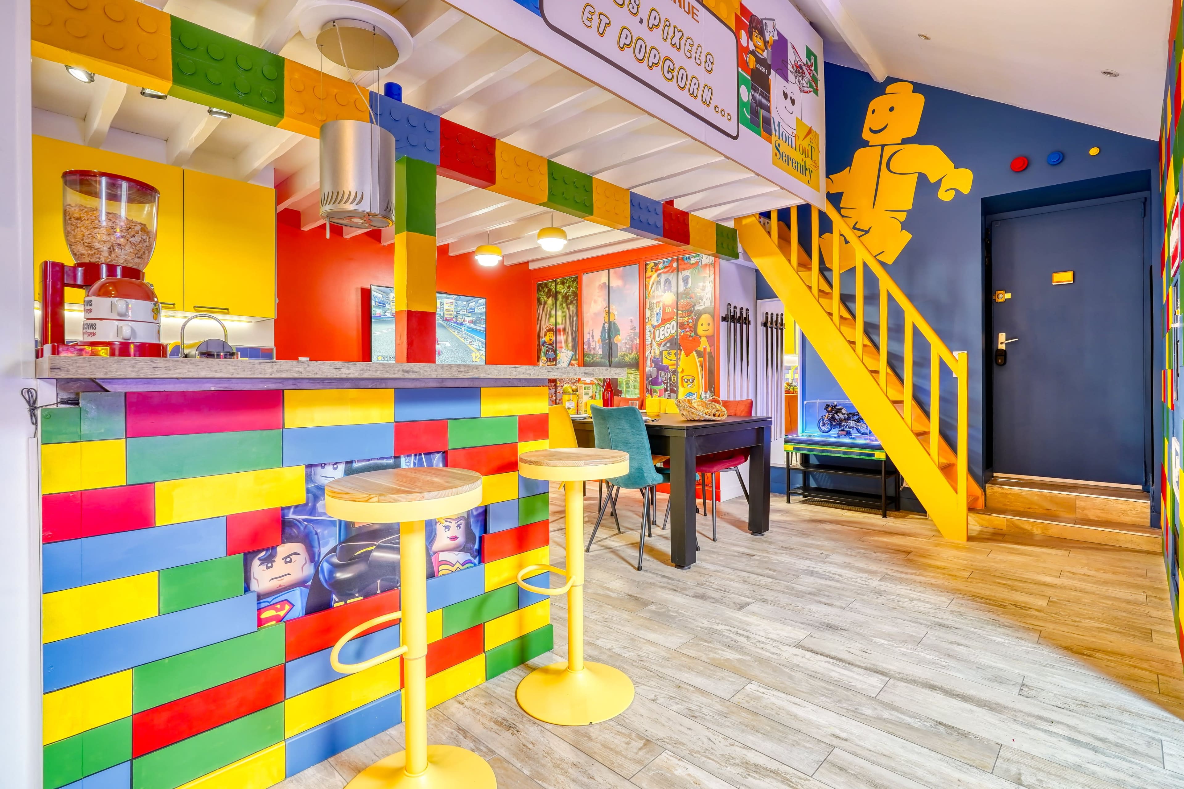The image shows a brightly colored interior, featuring a kitchen and dining area with walls and a bar decorated in a Lego theme, including yellow stools and a staircase.