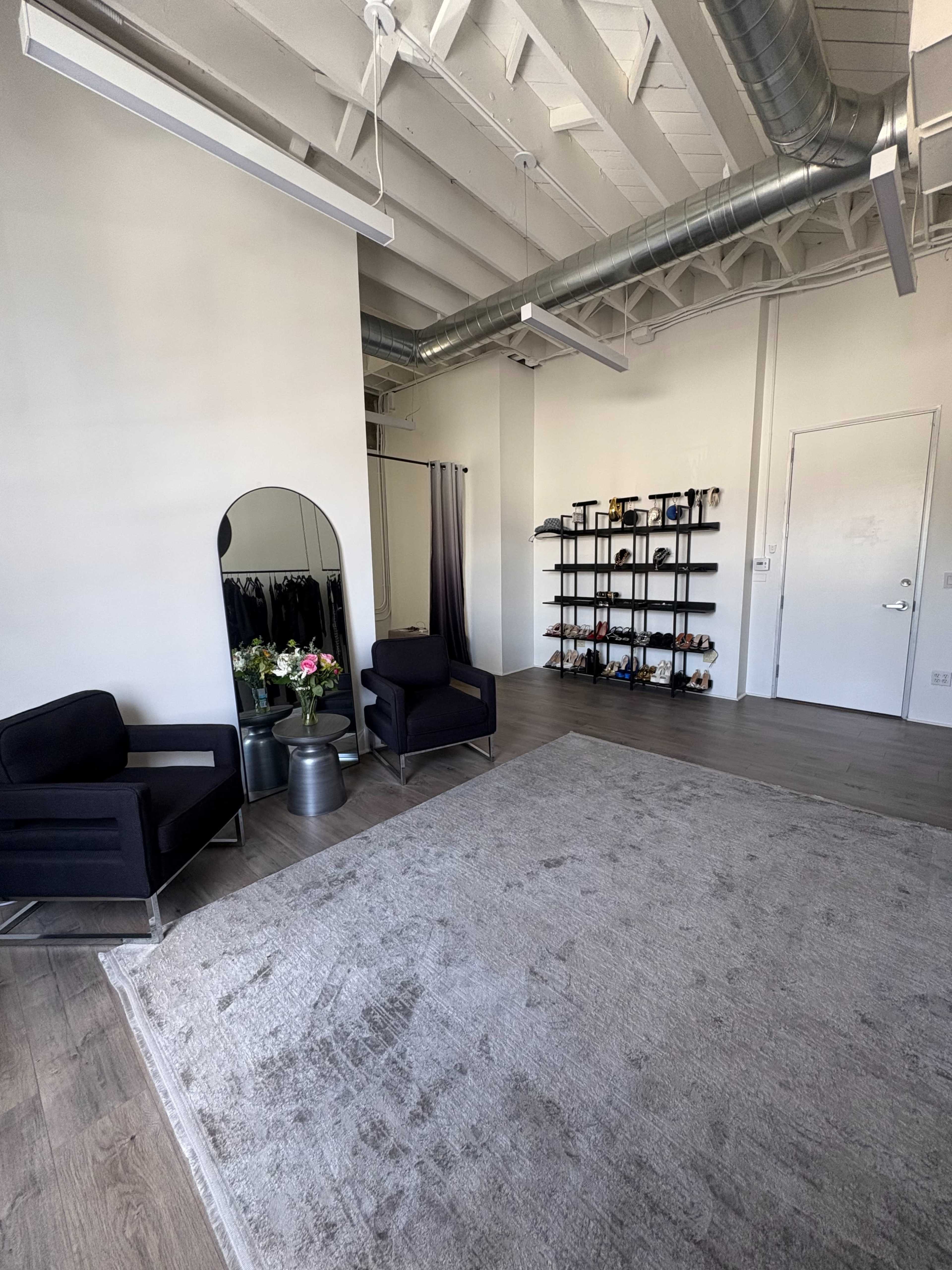 The image shows a minimalist room with two black chairs, a mirror, a shelf displaying shoes, and a floral arrangement on a small table.