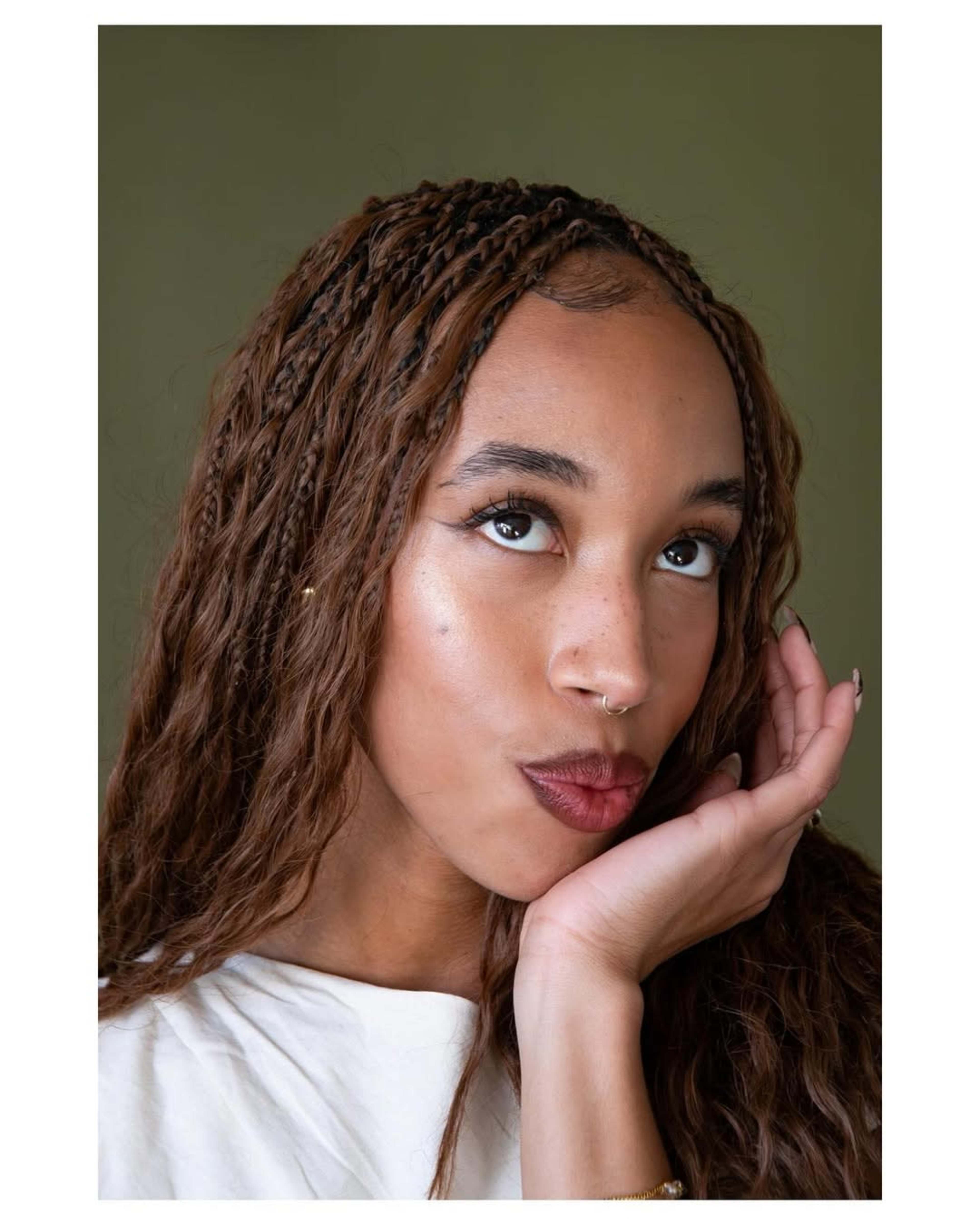 A person with braided hair poses with one hand on their cheek against a plain green background.