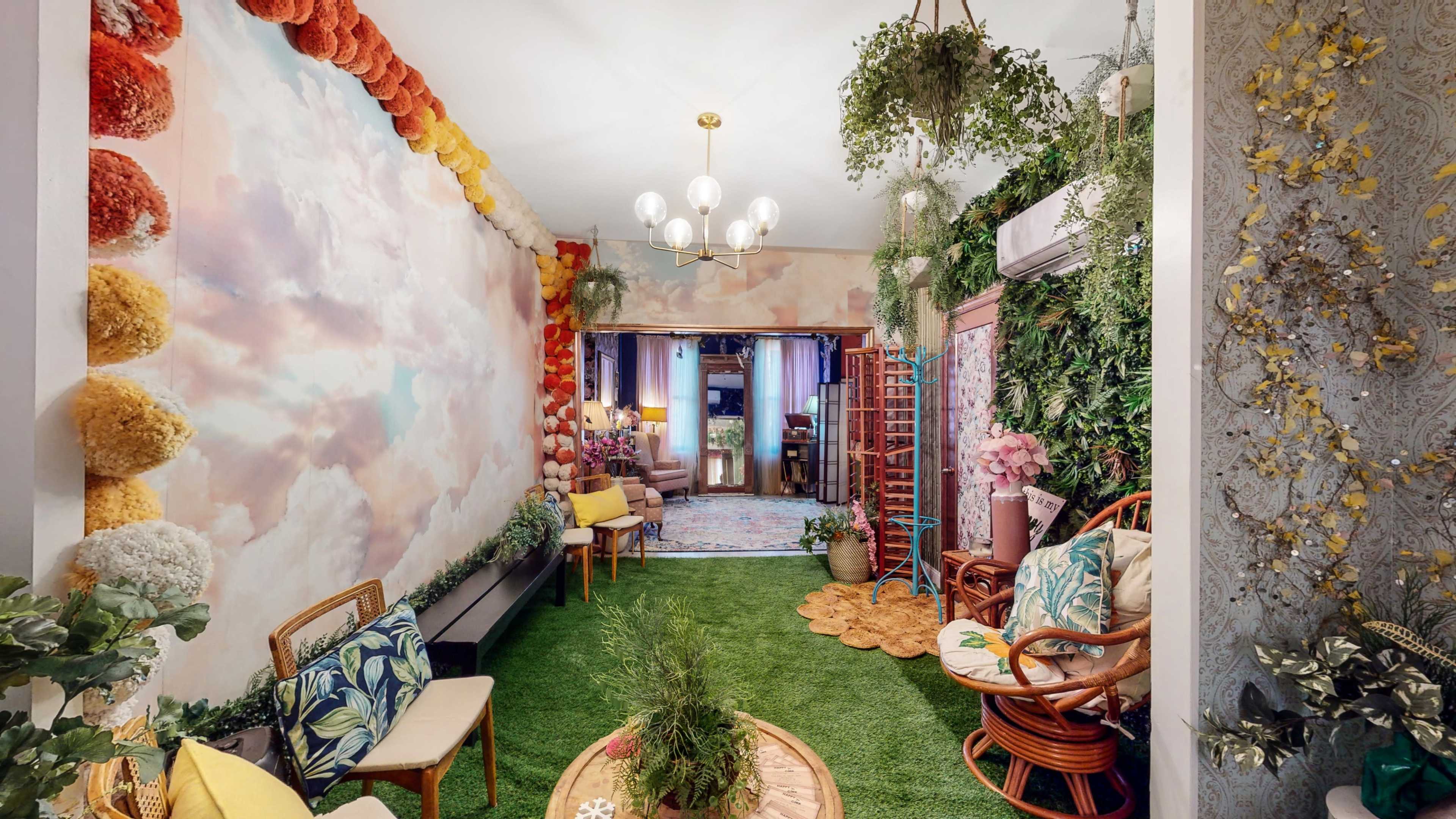 The scene depicts a vibrant indoor space with floral decor, greenery, and comfortable seating arranged around a circular rug.