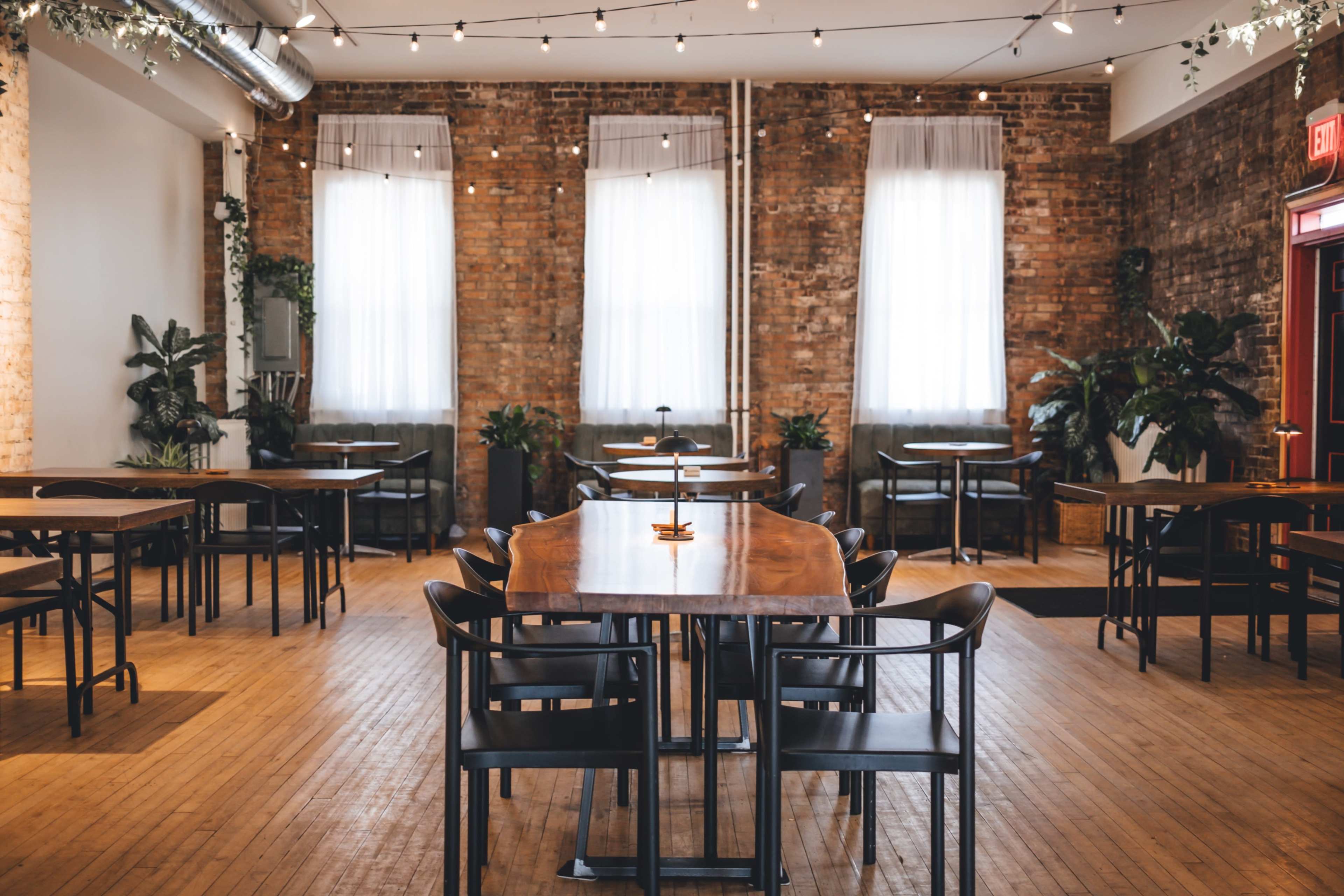 The image shows an indoor dining area with wooden tables and chairs, large windows, and plants against a brick wall.