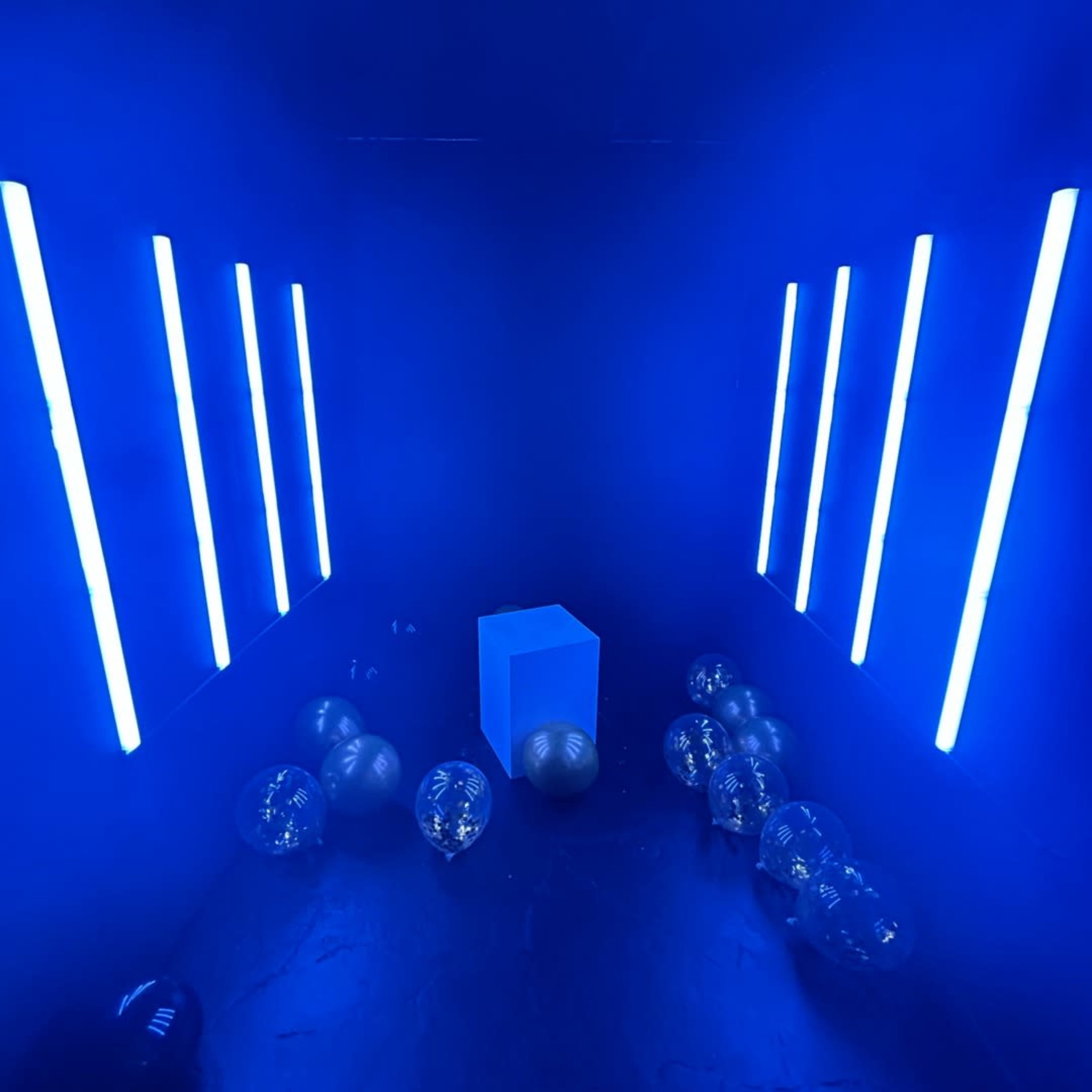 A dark room illuminated by blue lights with a blue cube in the center and transparent balloons scattered on the floor.