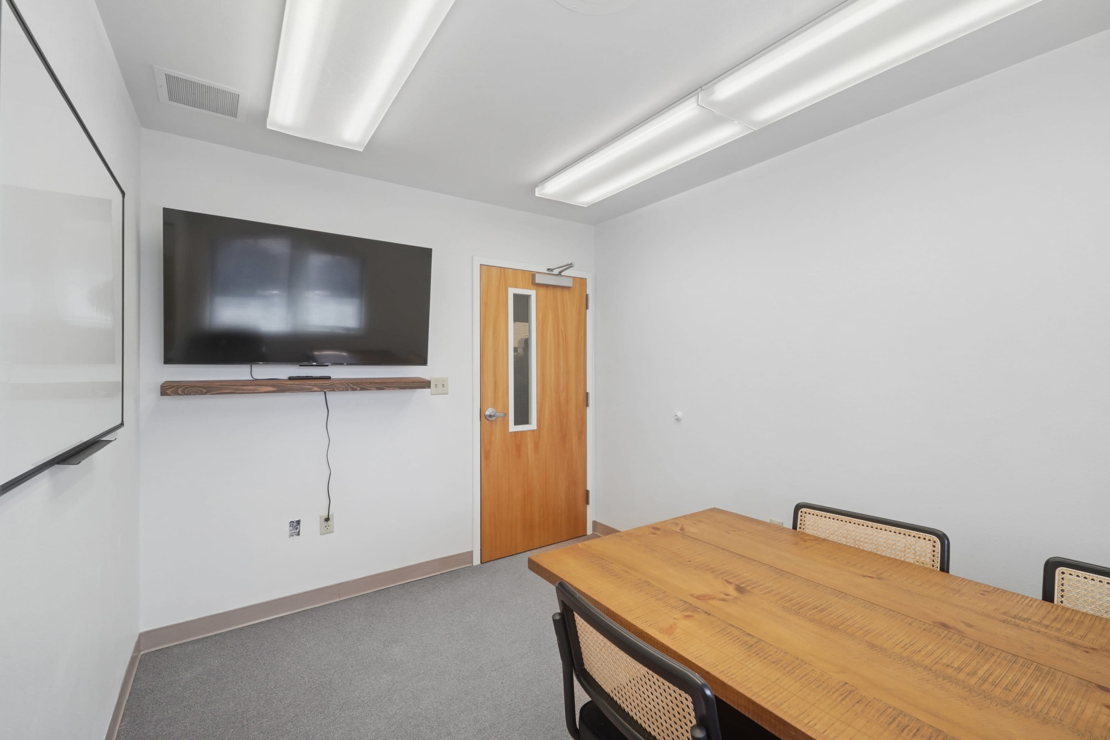 The image shows a small, sparsely furnished meeting room with a wooden table, chairs, a wall-mounted television, and a door with a small window.