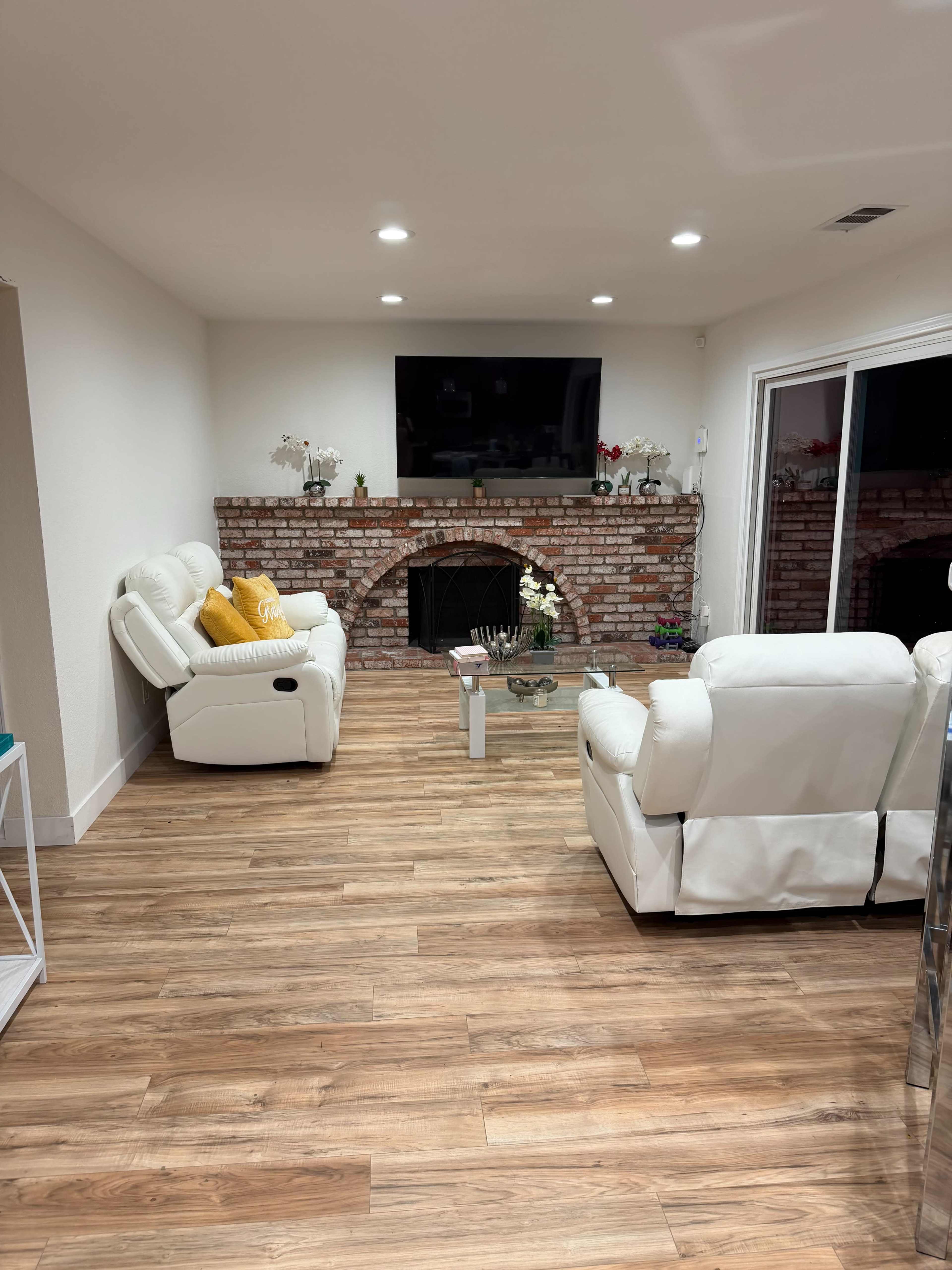 The image shows a living room with light-colored walls, a brick fireplace, two recliner chairs, a glass coffee table, and a large TV mounted on the wall.
