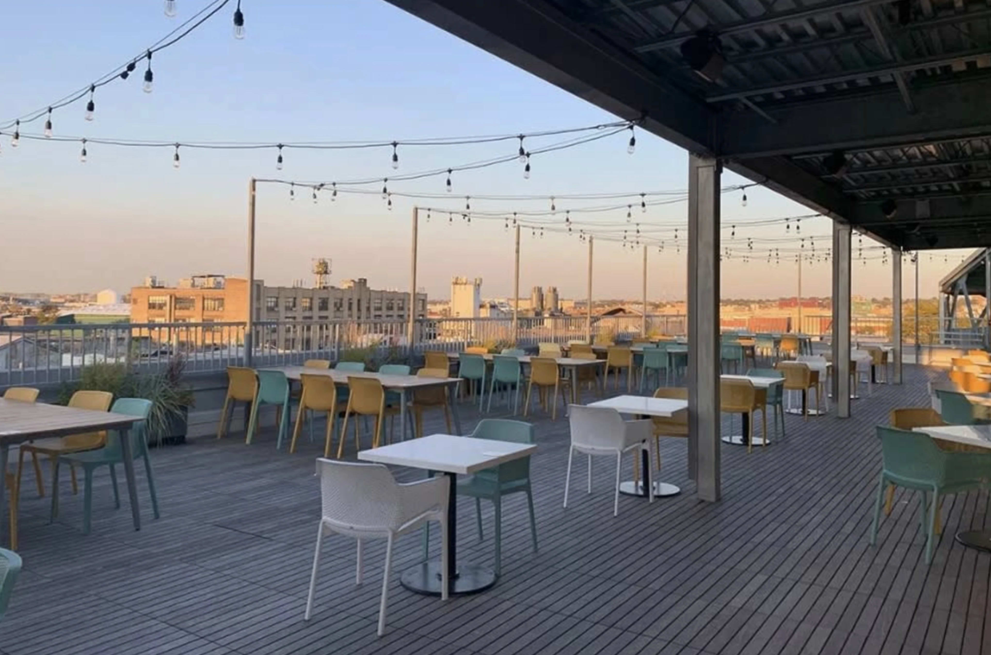 The image shows a rooftop terrace with wooden flooring, tables, and chairs arranged under string lights, with buildings visible in the background.