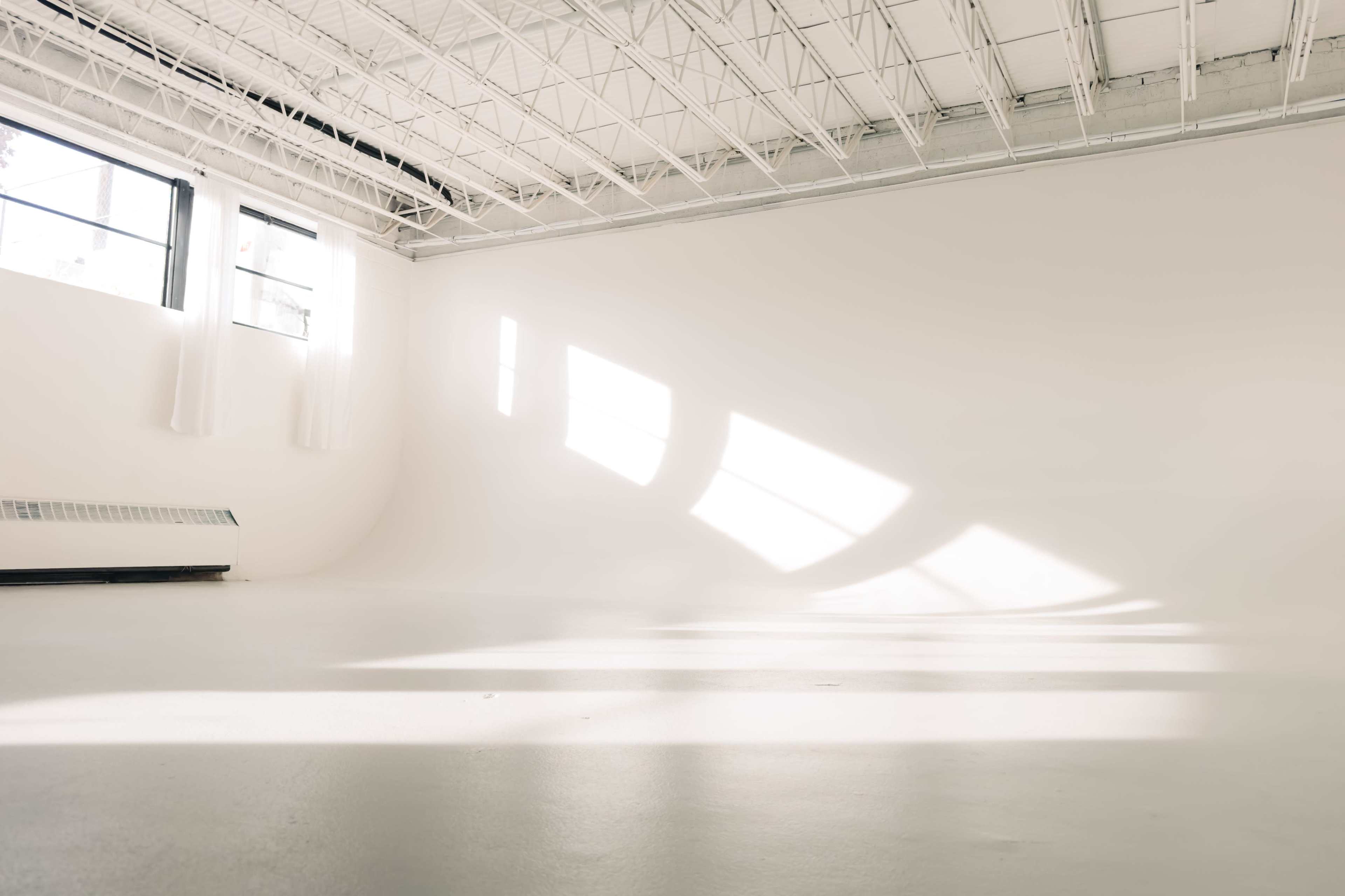 The image shows a spacious, brightly lit room with large windows and smooth white walls and floors.