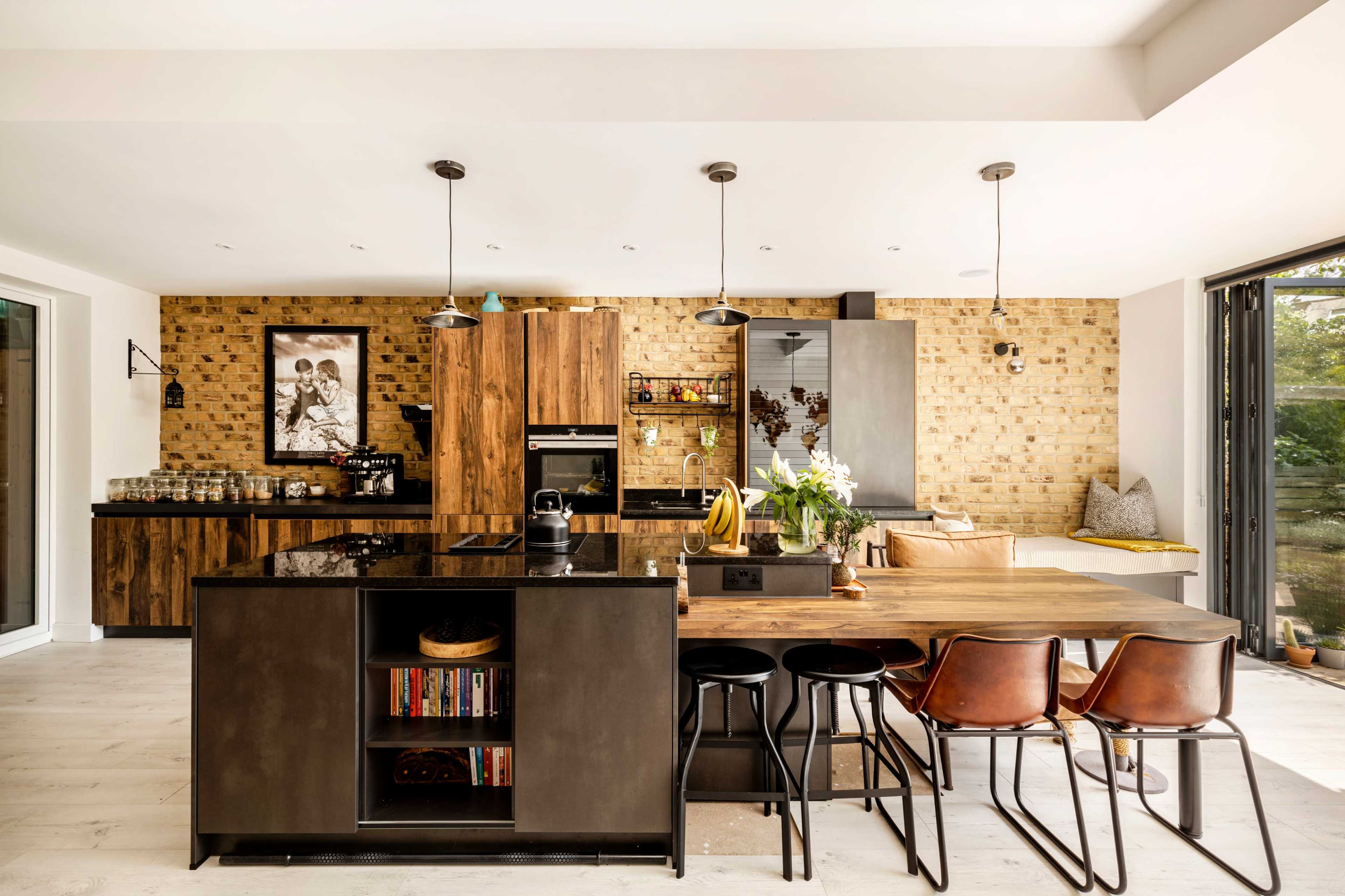 A modern kitchen features wooden cabinets, a central island, and a dining table with leather chairs, accented by a brick wall and large windows.