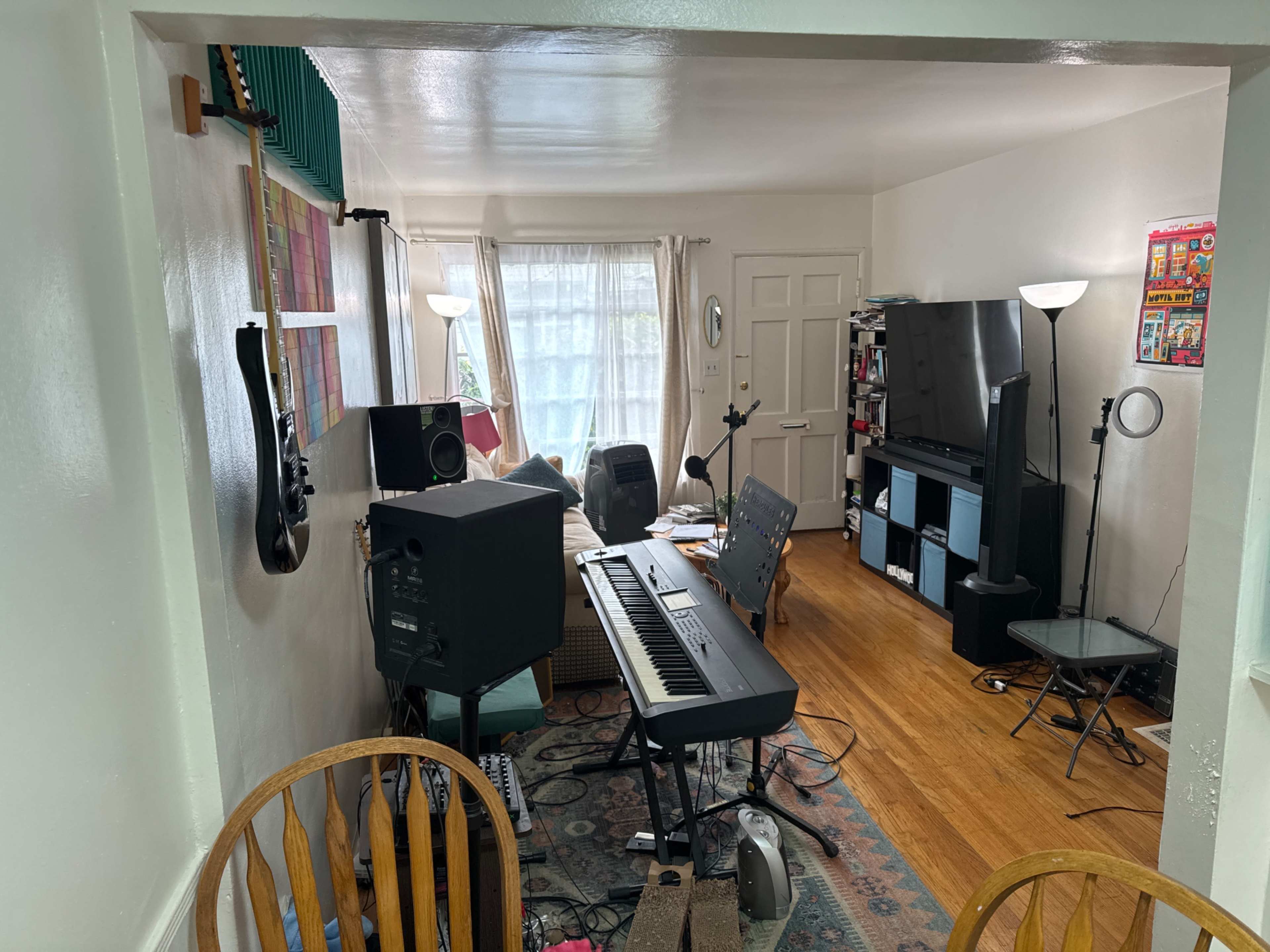 A cluttered living space features a keyboard, speakers, a television, and various furniture, illuminated by natural light coming through a window.