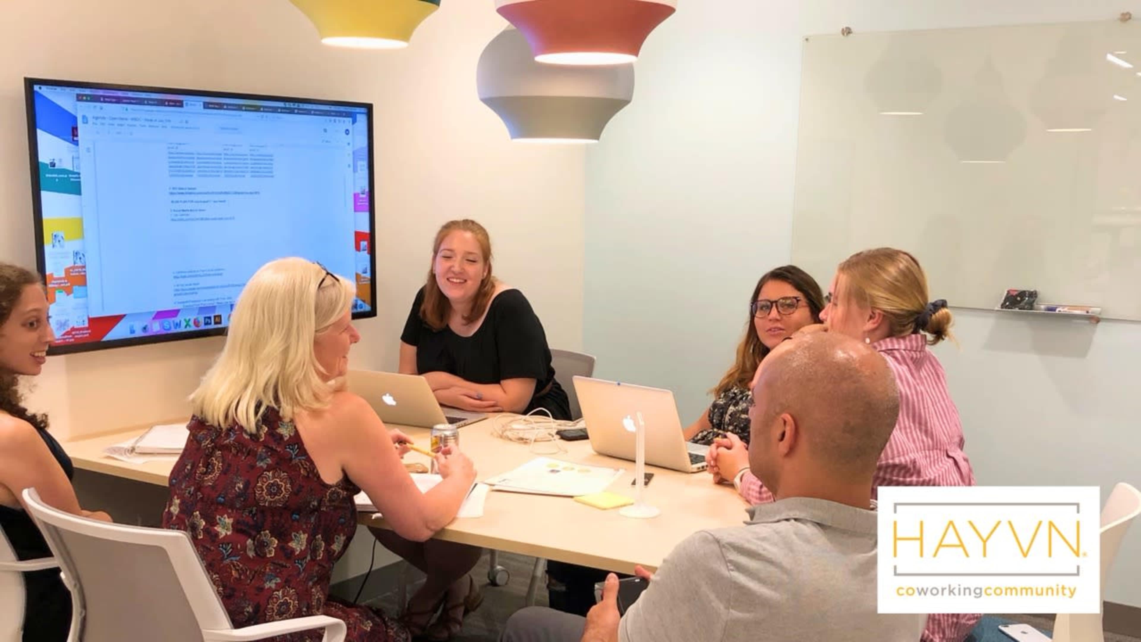 A group of six people is sitting around a table in a bright conference room, engaged in a discussion with laptops and documents in front of them.