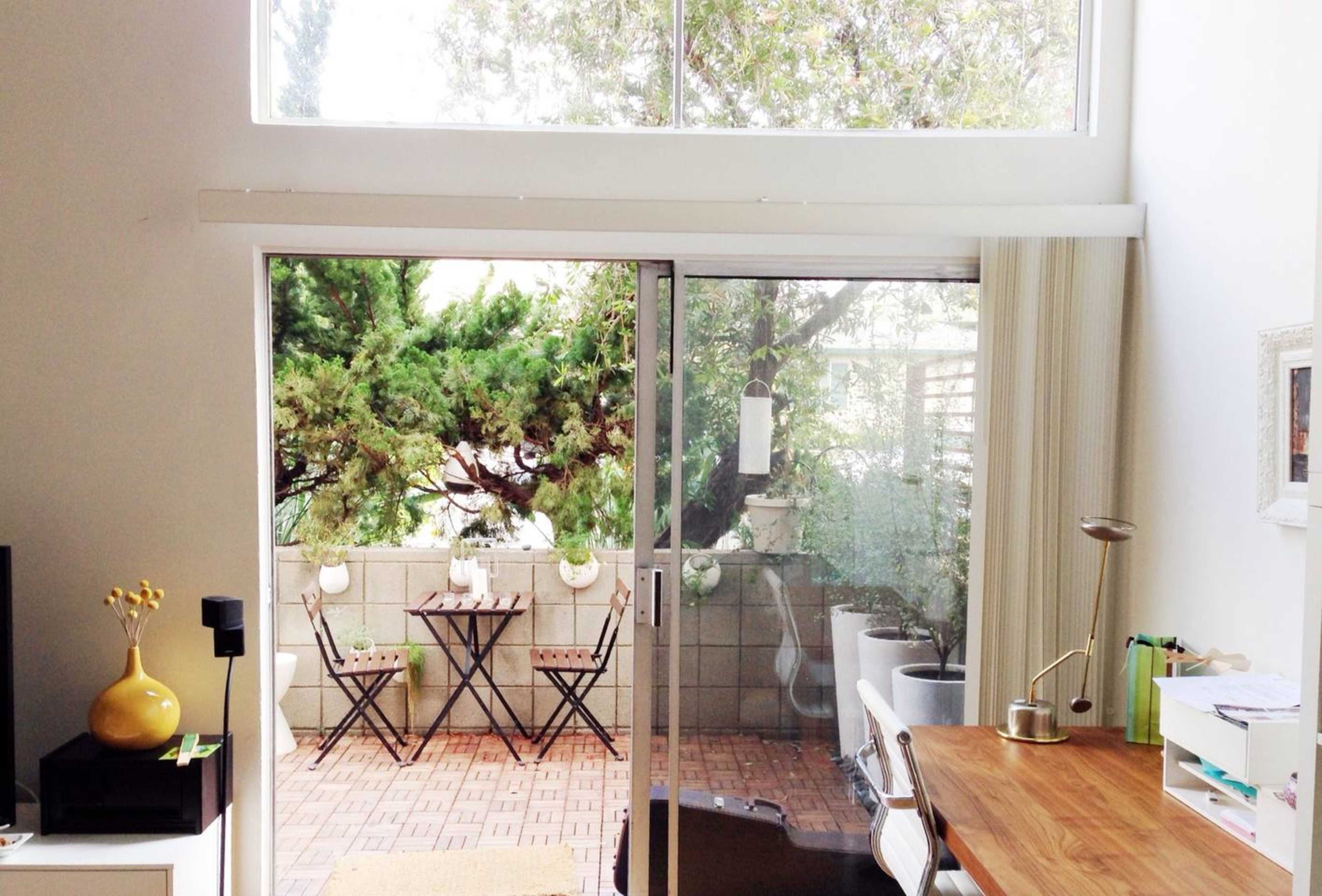 A bright room features a sliding glass door leading to a small patio with two chairs and a table, surrounded by greenery.