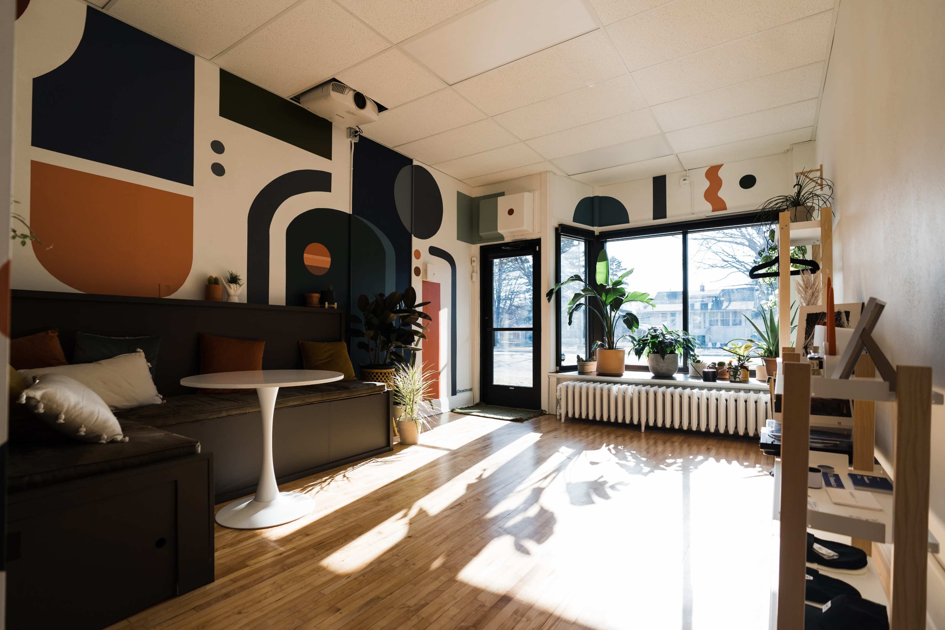 A modern interior space with a patterned mural on the wall, a round table, seating area, and large windows allowing natural light to enter.