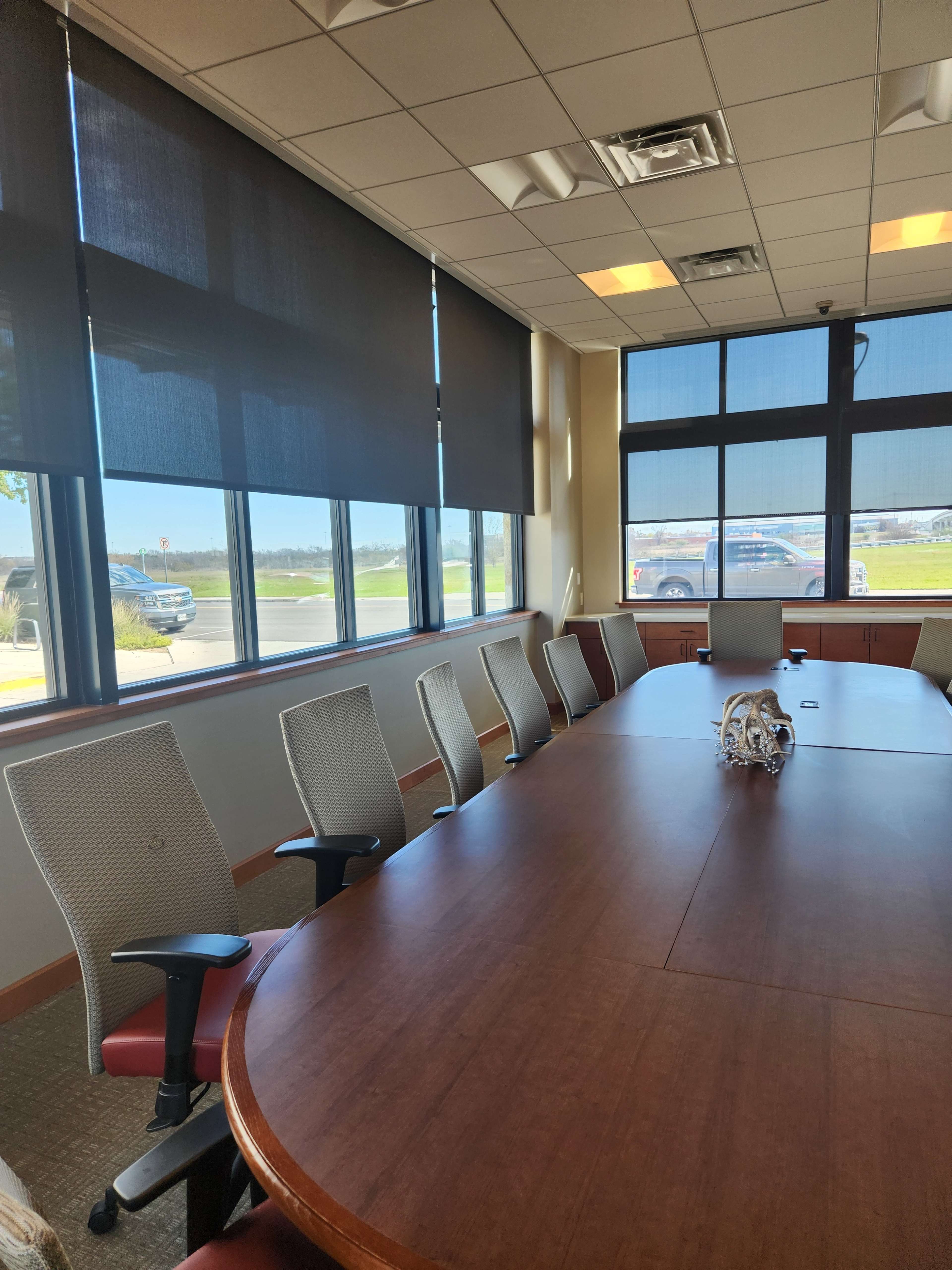 A large conference room features a rectangular wooden table surrounded by eight office chairs, with large windows letting in natural light.