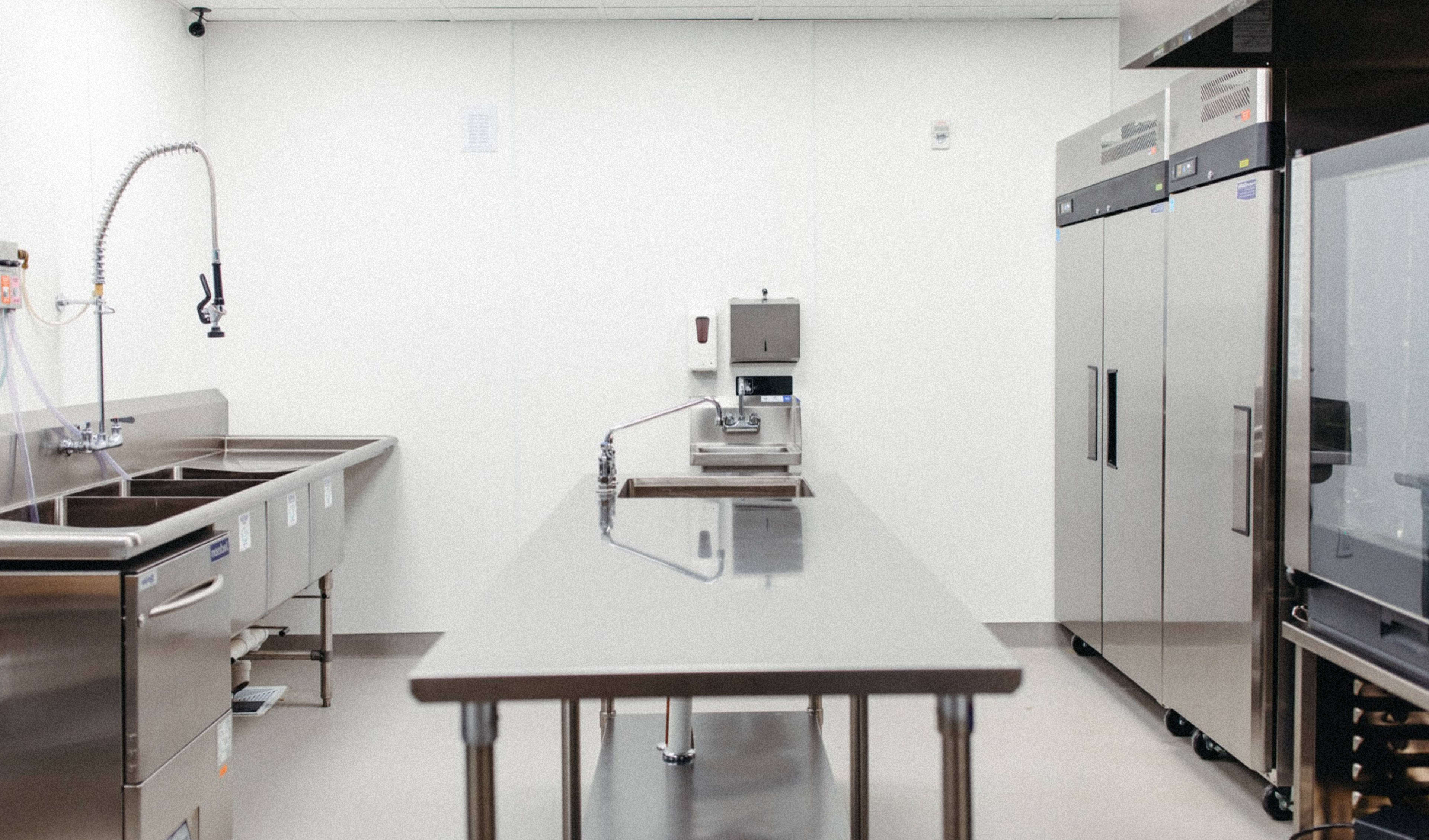 A clean, stainless steel kitchen workspace features a table in the center, sinks on one side, and refrigerators on the other.