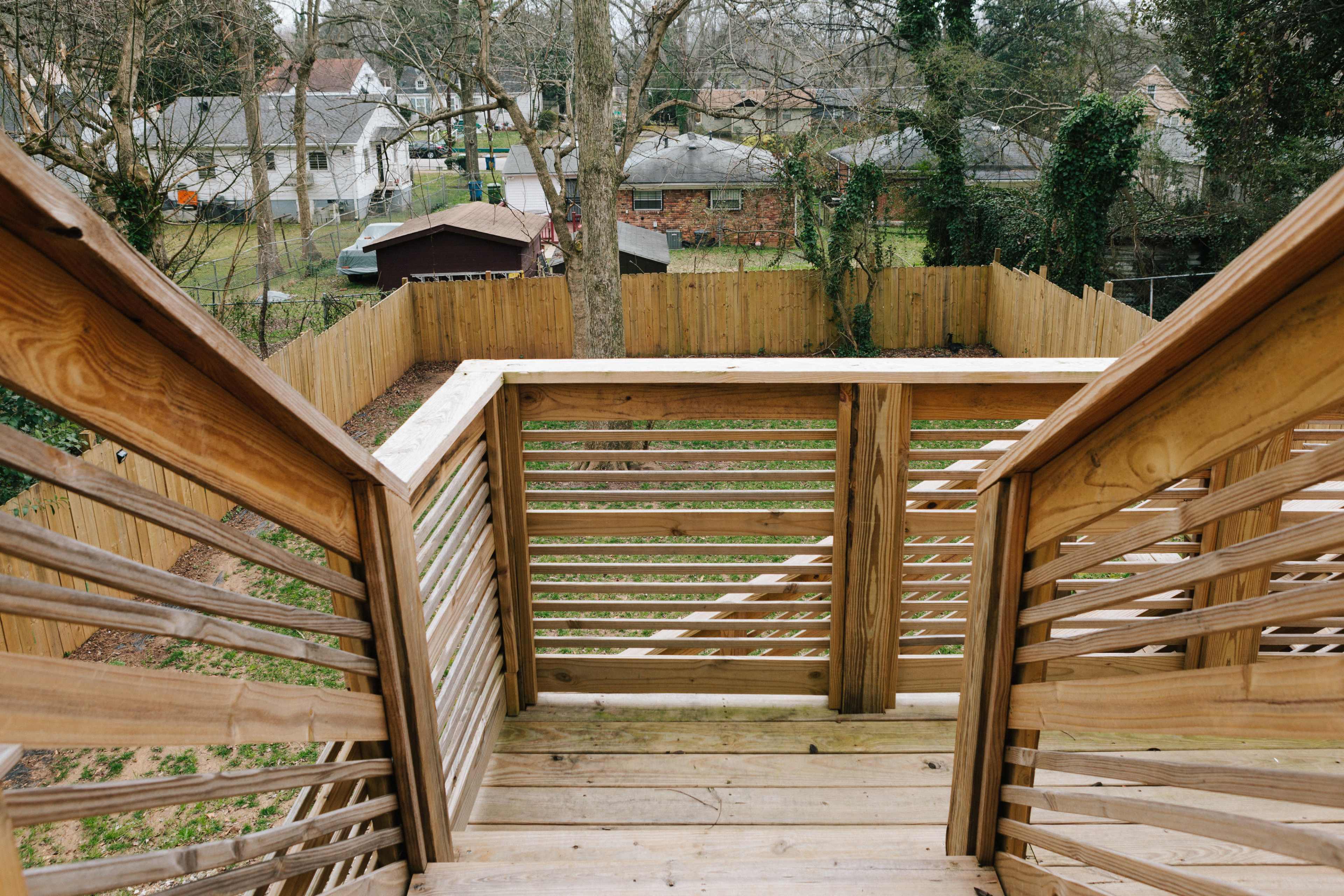 A wooden staircase leading down to a backyard area surrounded by a wooden fence and several trees and buildings in the distance.