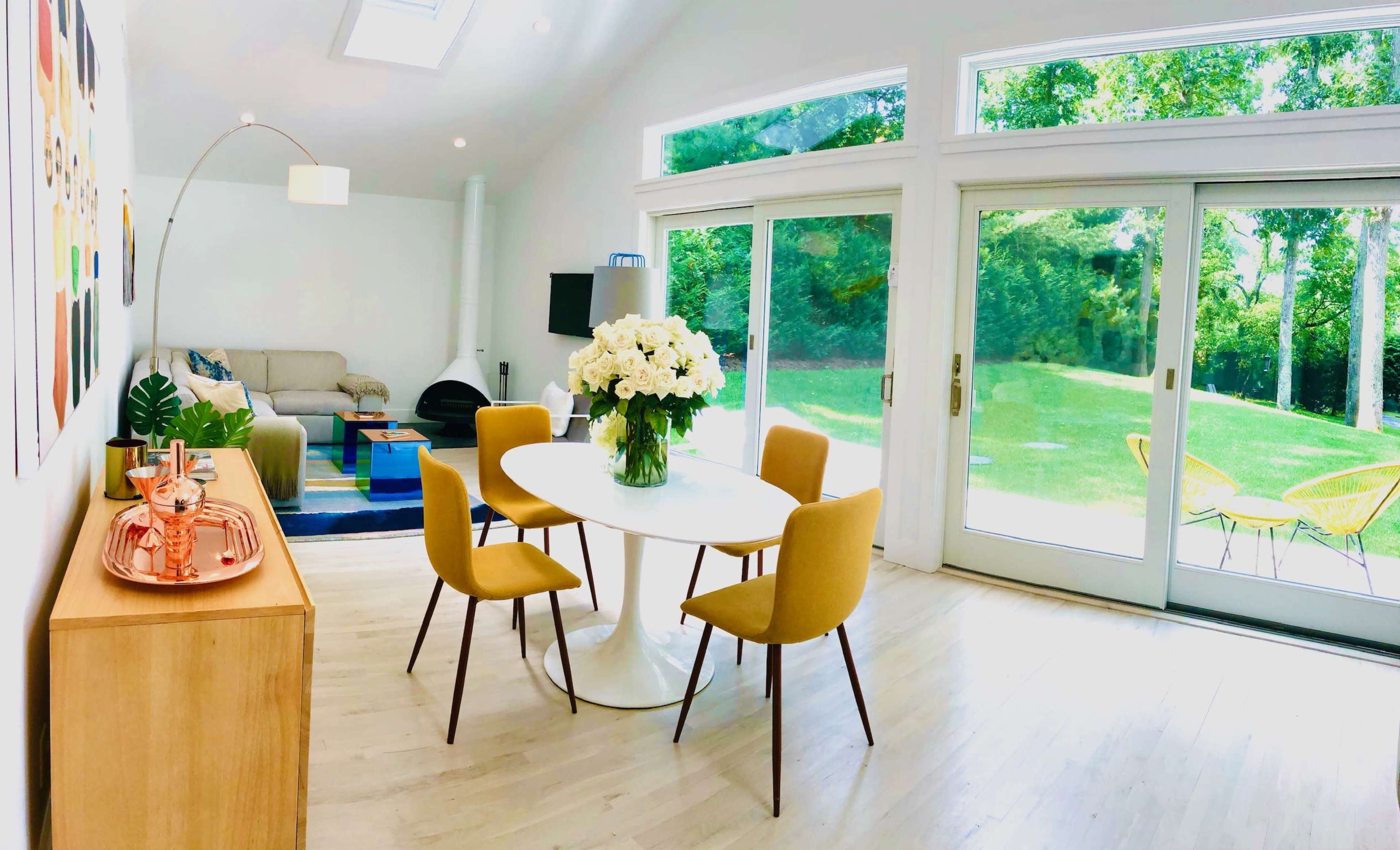 The image shows a bright, modern living space with a round dining table surrounded by four yellow chairs, a vase of white flowers on the table, and large glass doors opening to a grassy outdoor area.